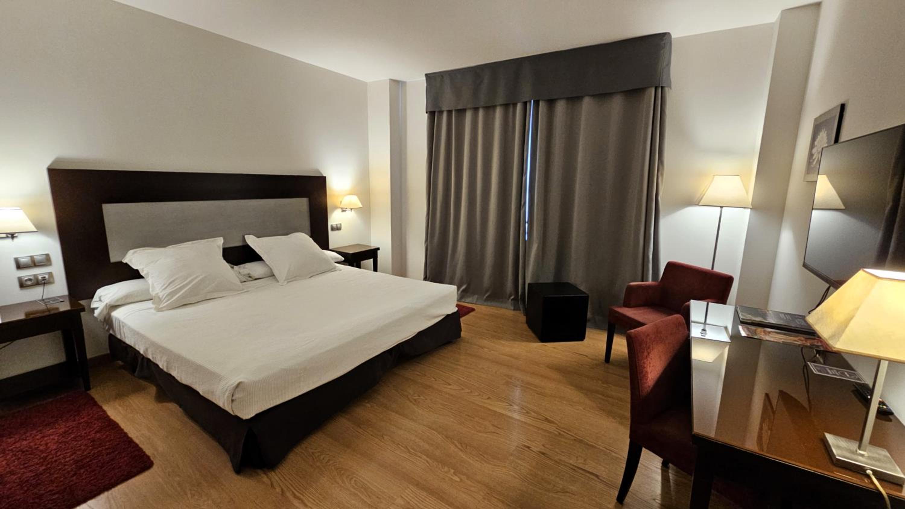 A hotel room at Las Bóvedas with a large made bed, dark headboard, wood flooring, red chairs, and a desk.