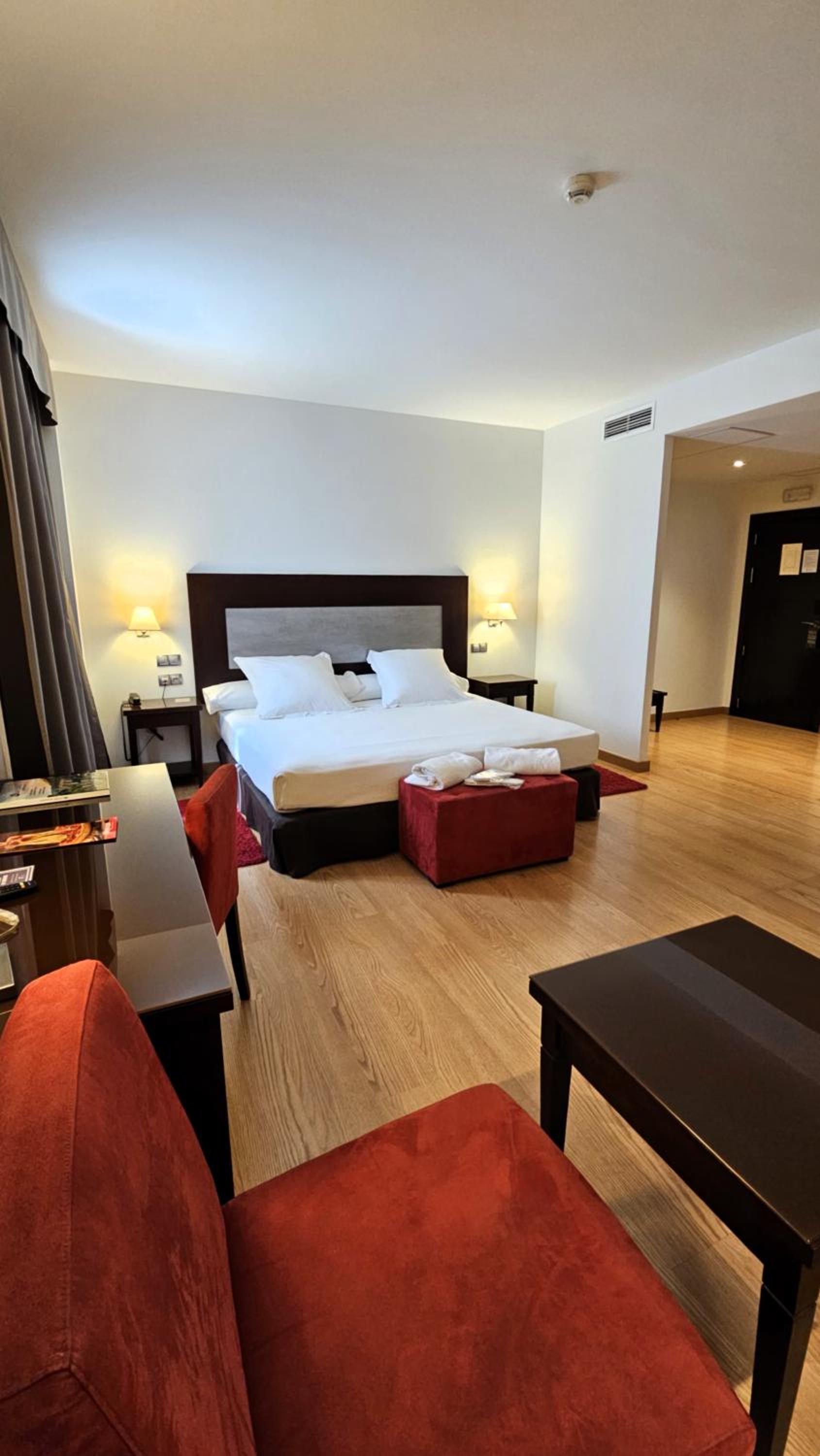 A hotel bedroom featuring wood flooring, red upholstered chairs, and a large bed with a dark headboard.
