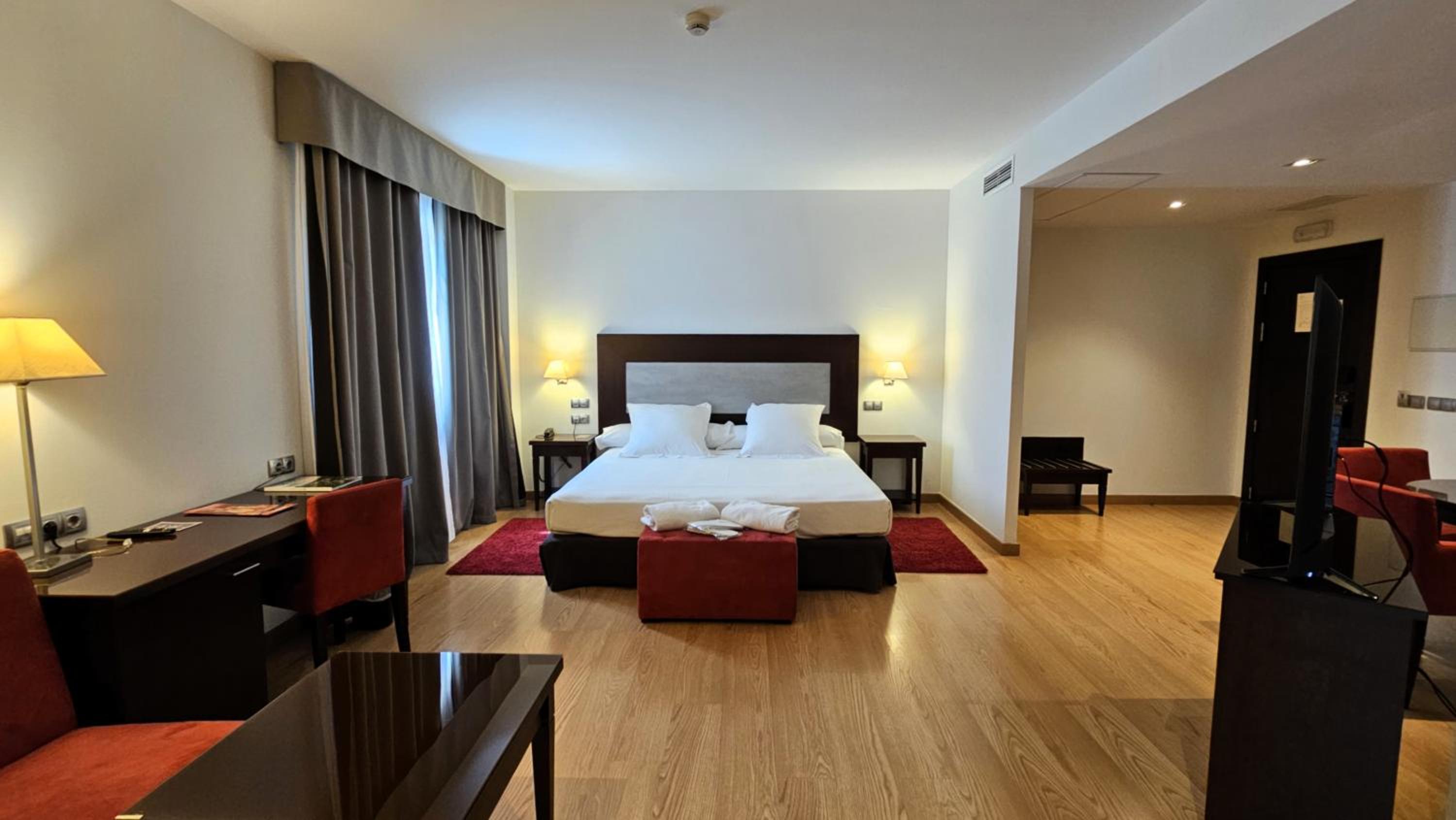 A hotel bedroom with a large bed, matching nightstands, red rugs, and a work desk.