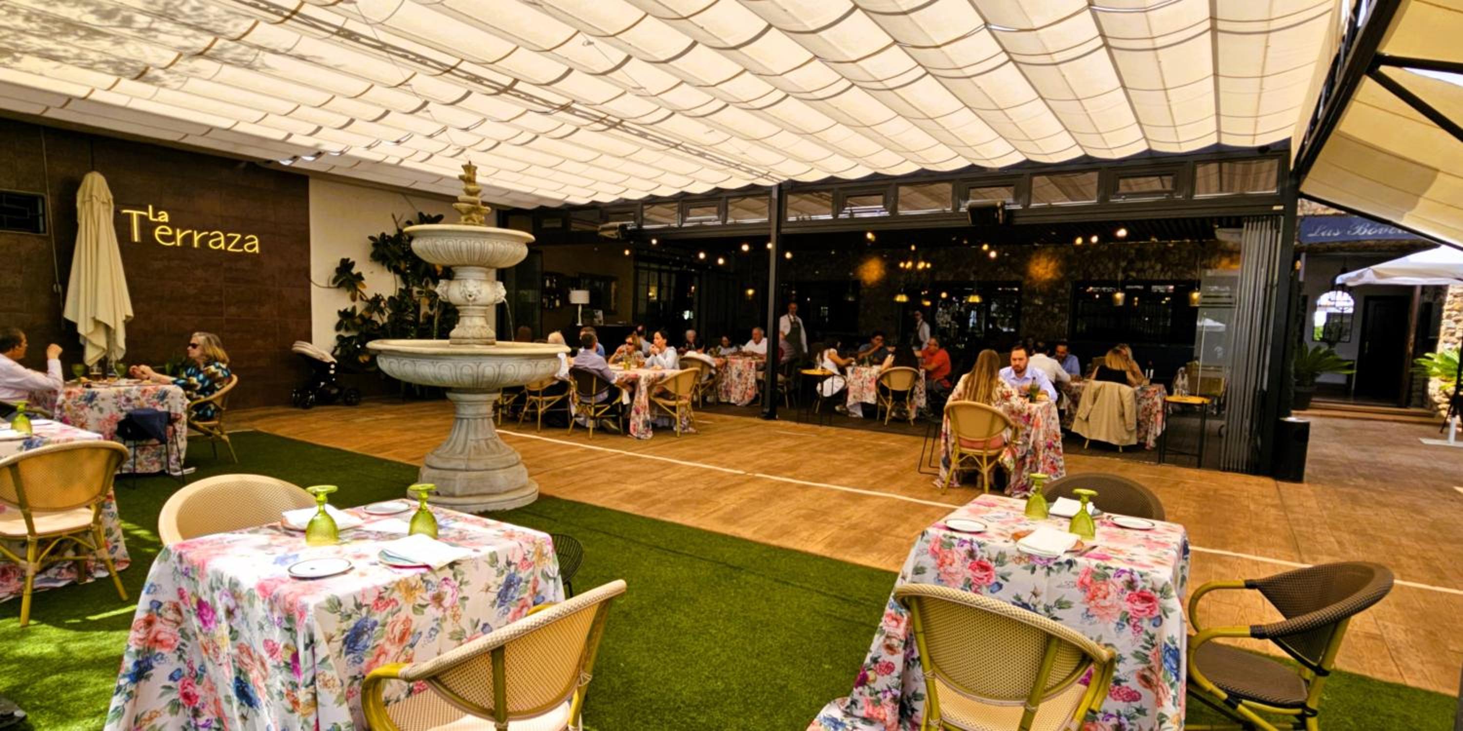 A covered outdoor terrace dining area with a stone fountain, tables dressed in floral cloths, and artificial grass.