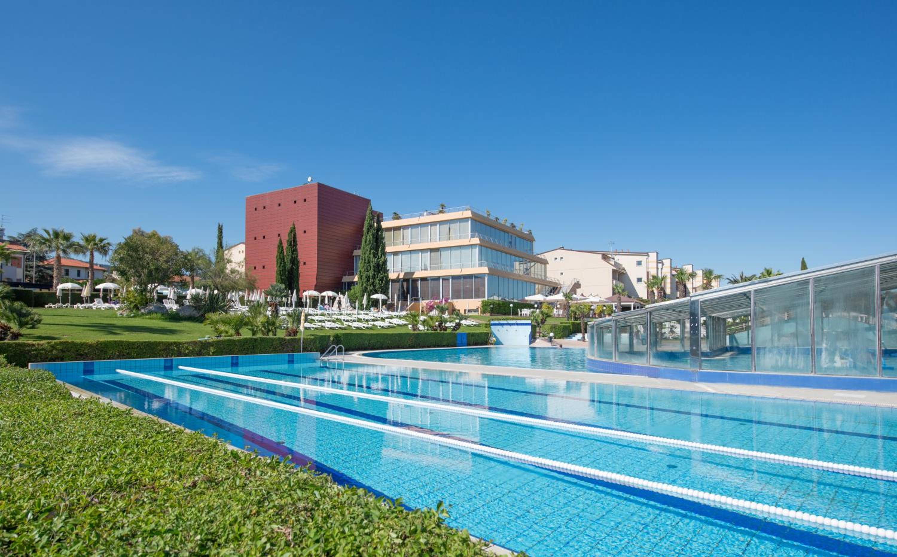 An outdoor swimming pool with lane markers sits beside a lawn leading to modern resort buildings.