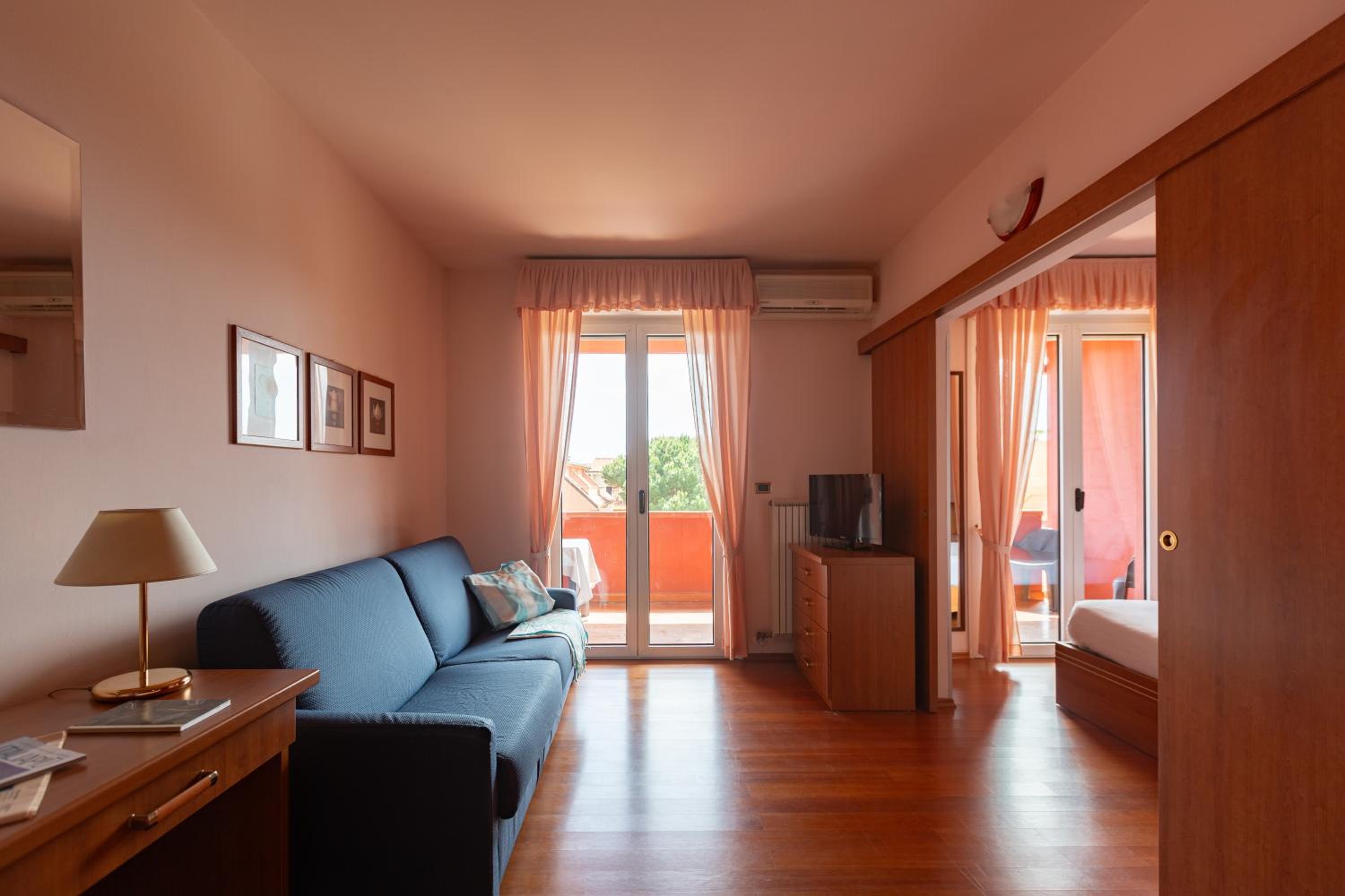 A hotel living area features a blue sofa, polished wood flooring, and sheer curtains framing glass balcony doors.