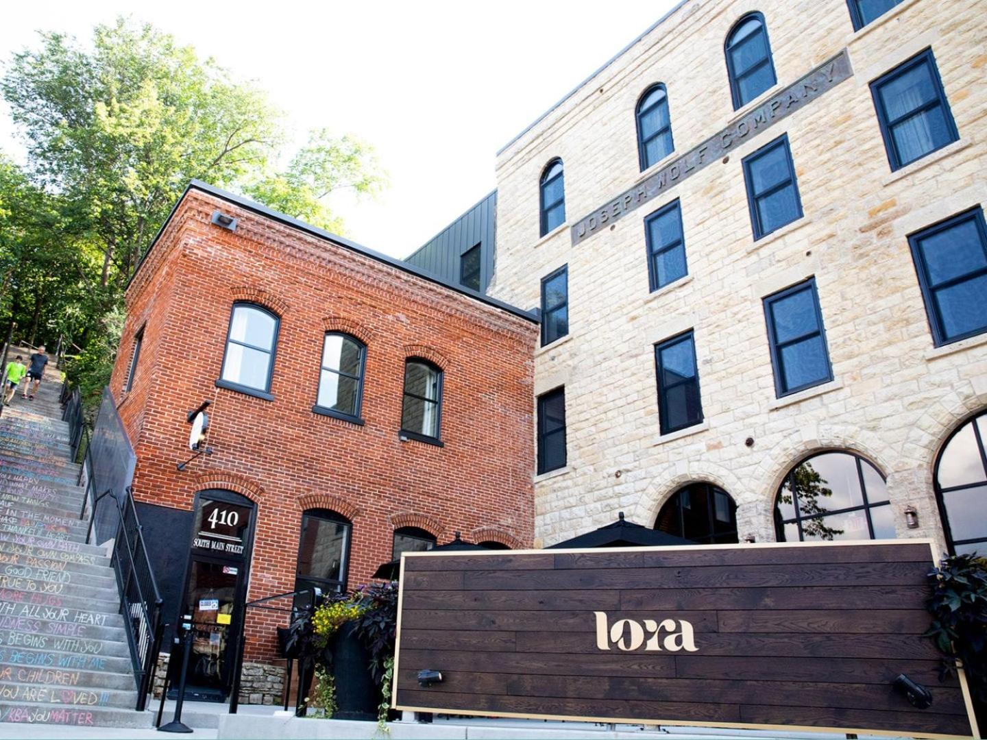 Brick and stone building facades rise beside an outdoor staircase, fronted by a dark wooden sign for Lora.