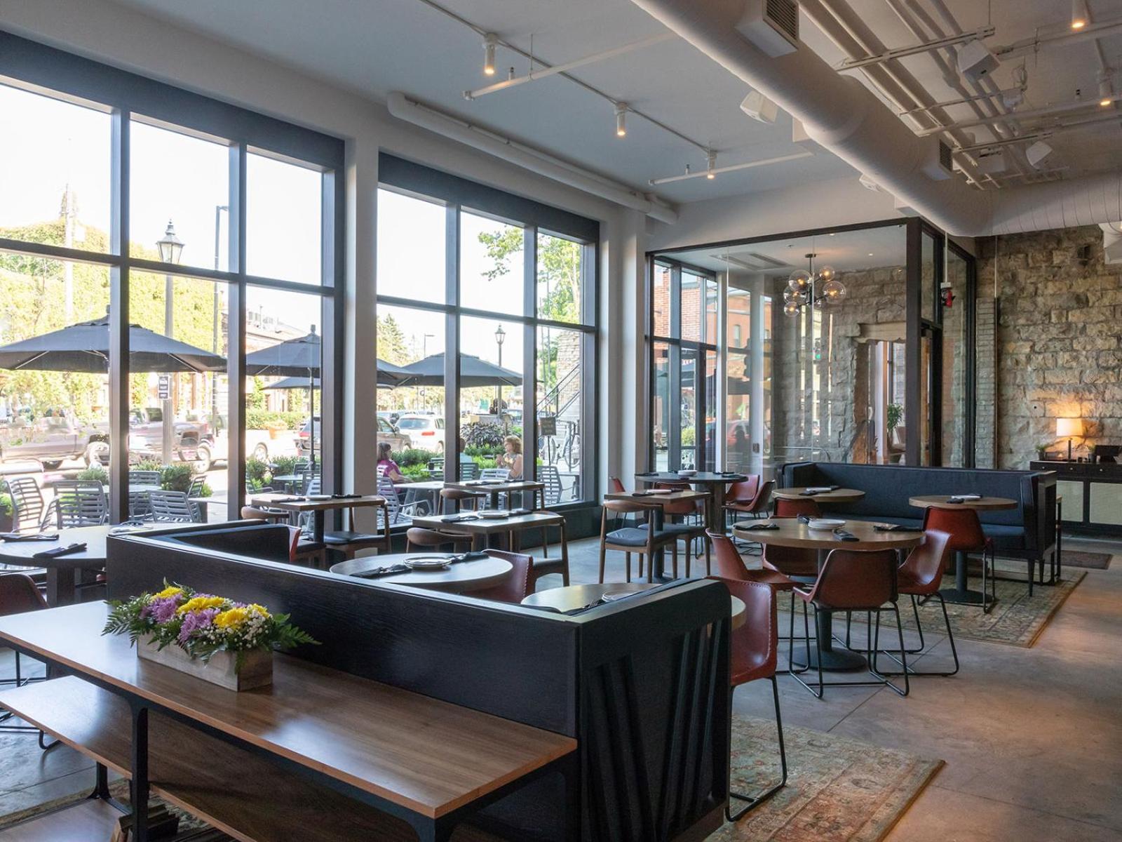 A sunlit dining room features large black-framed windows looking out onto a patio with umbrella-shaded tables.