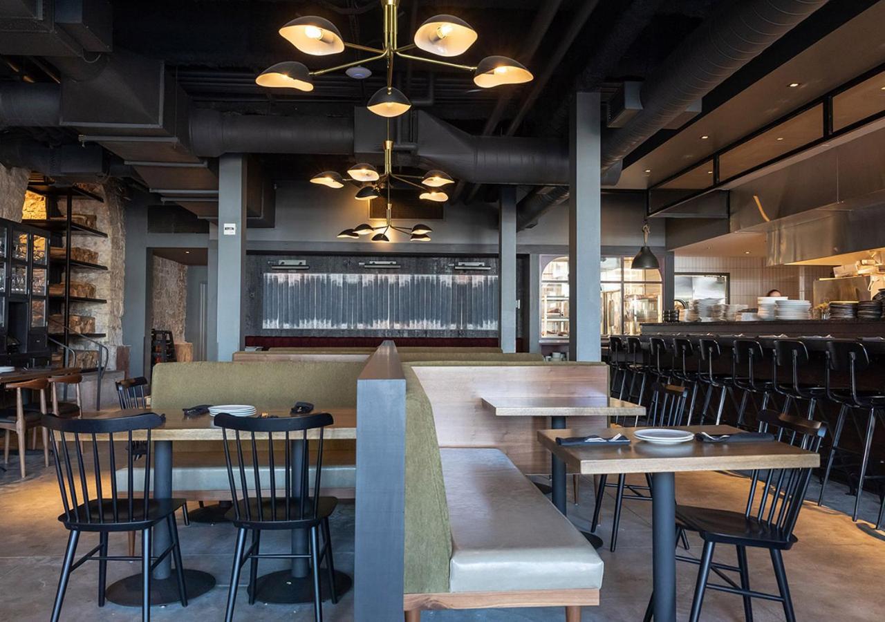 Dining room interior with upholstered banquettes, dark exposed ductwork, brass chandeliers, and an open kitchen counter.