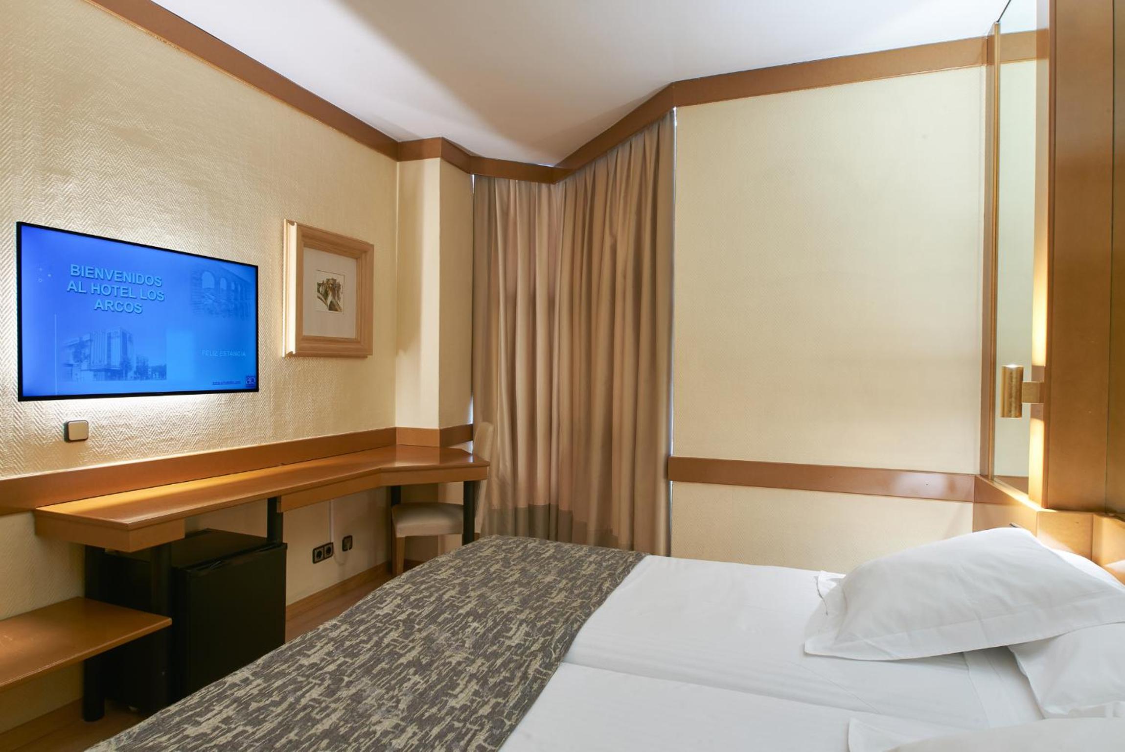 Corner of a hotel room with a television mounted above a desk, a mini fridge underneath, and curtained windows.