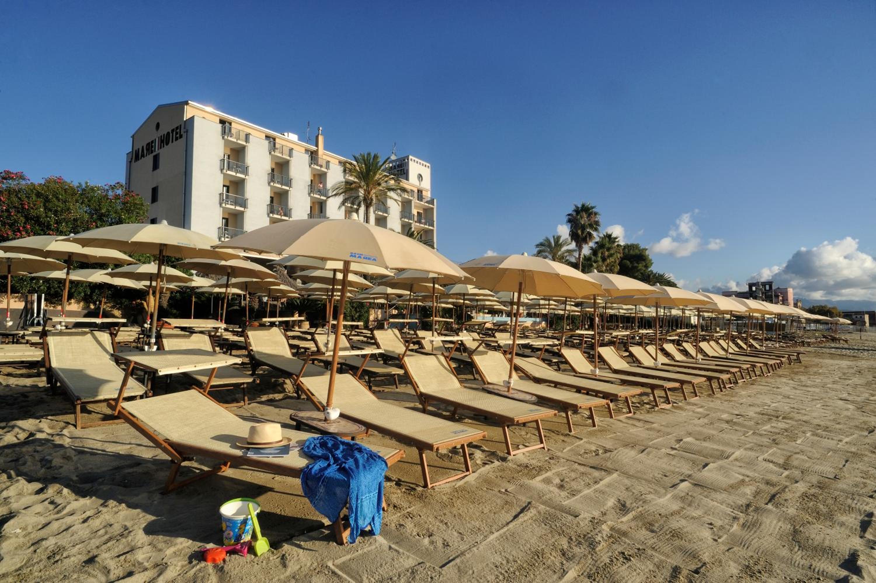 Sun loungers and umbrellas line a sandy beach in front of the Mare Hotel building under clear blue skies.