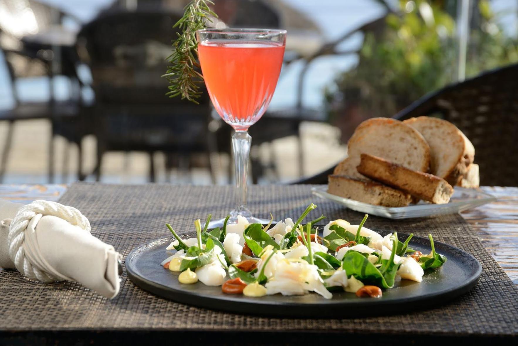 A seafood salad, sliced bread, and a pink cocktail garnished with rosemary sit on a woven outdoor table.