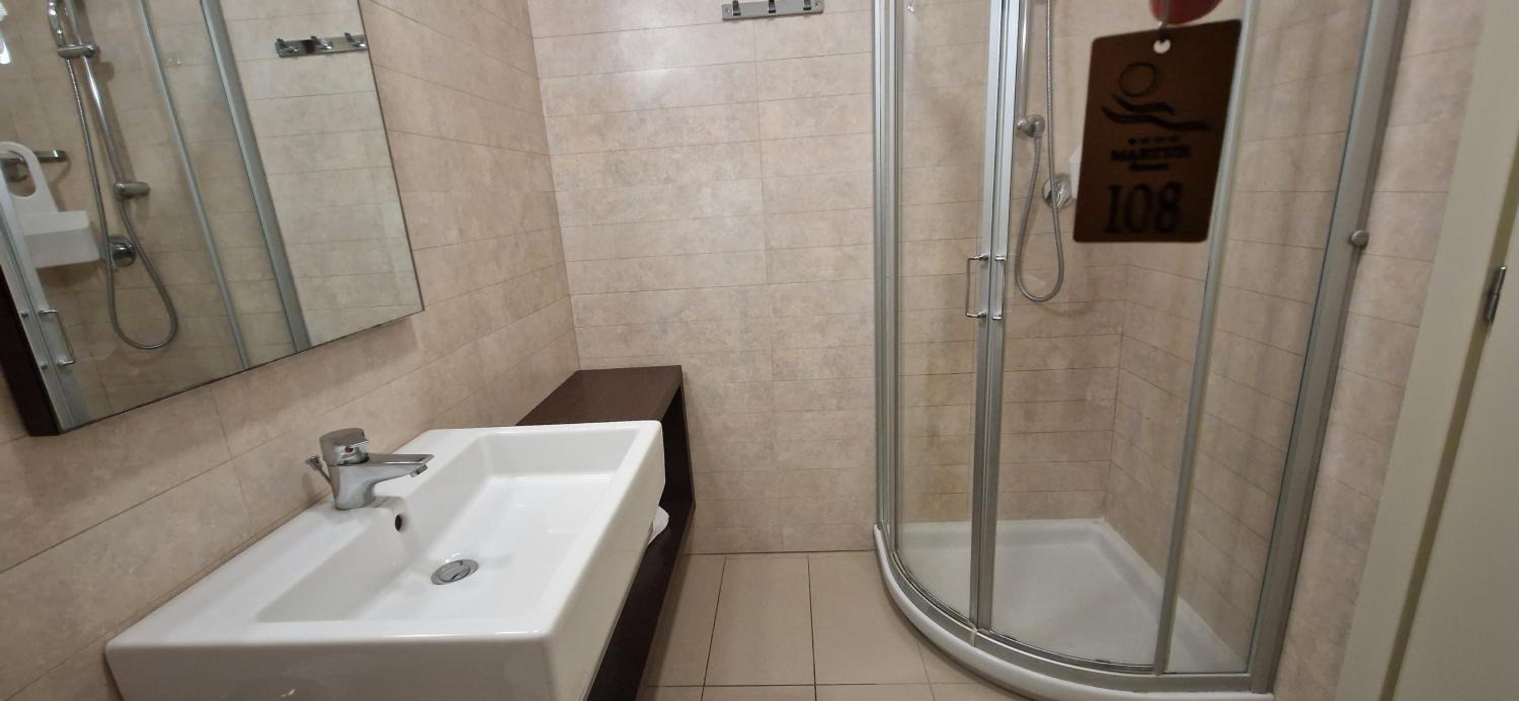 A modern bathroom with a white sink, glass corner shower stall, and light beige tiled walls.