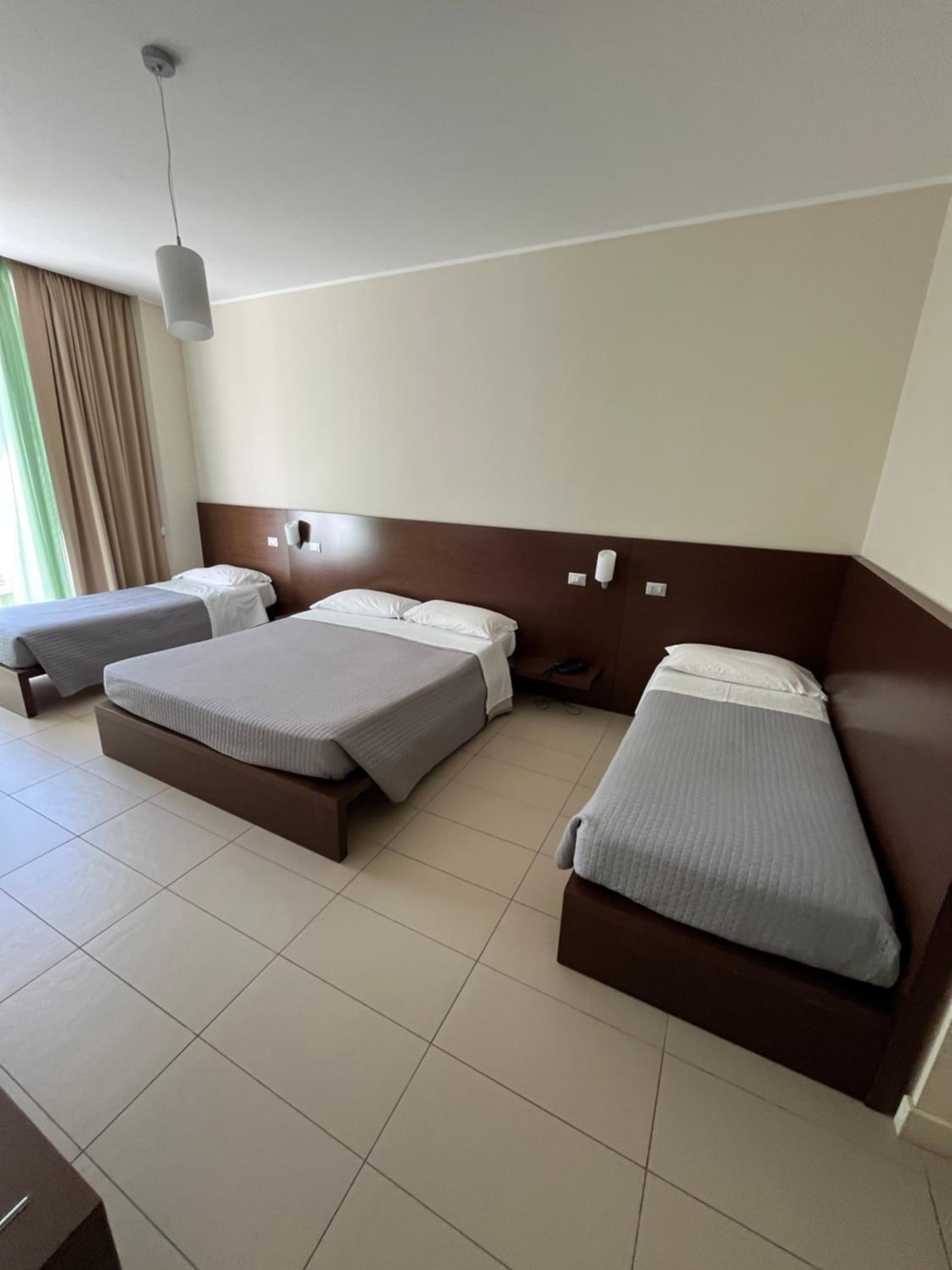 A hotel bedroom with tiled floors, a dark wood headboard, made beds, and neutral curtains.