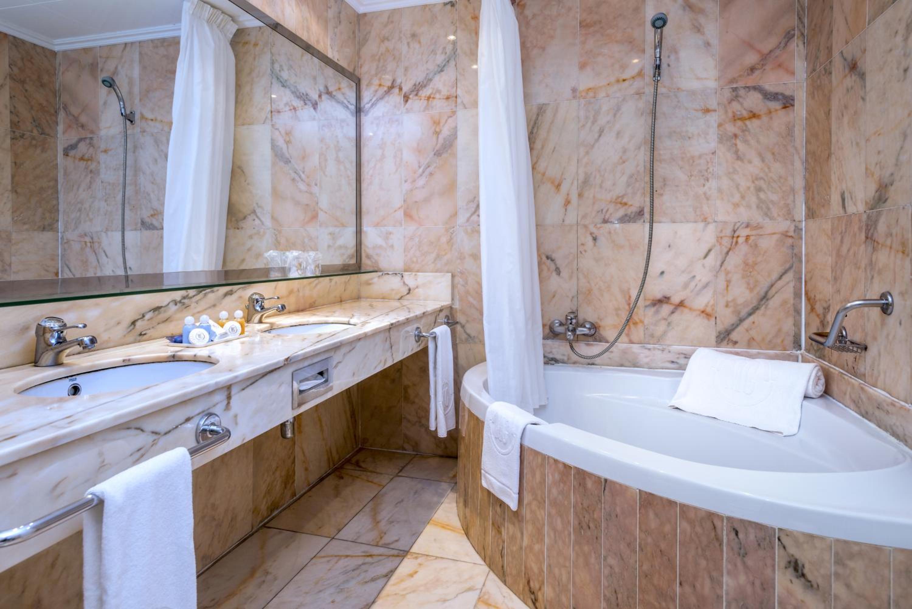 A marble-tiled bathroom with a vanity, sinks, a large mirror, and a curved bathtub with a shower curtain.
