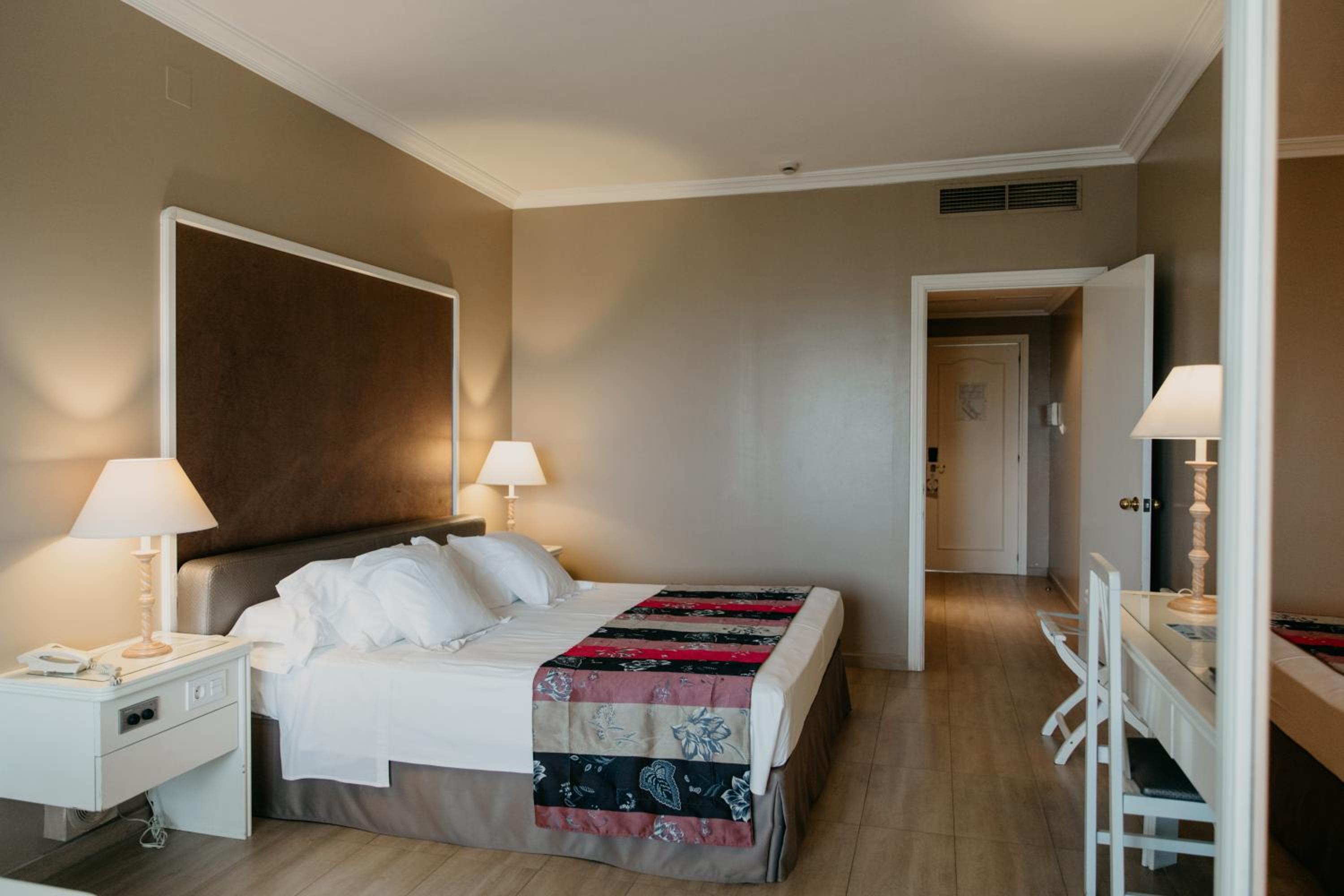 A hotel bedroom at Mas Gallau with a made bed, bedside lamps, neutral walls, and an open doorway to the hall.