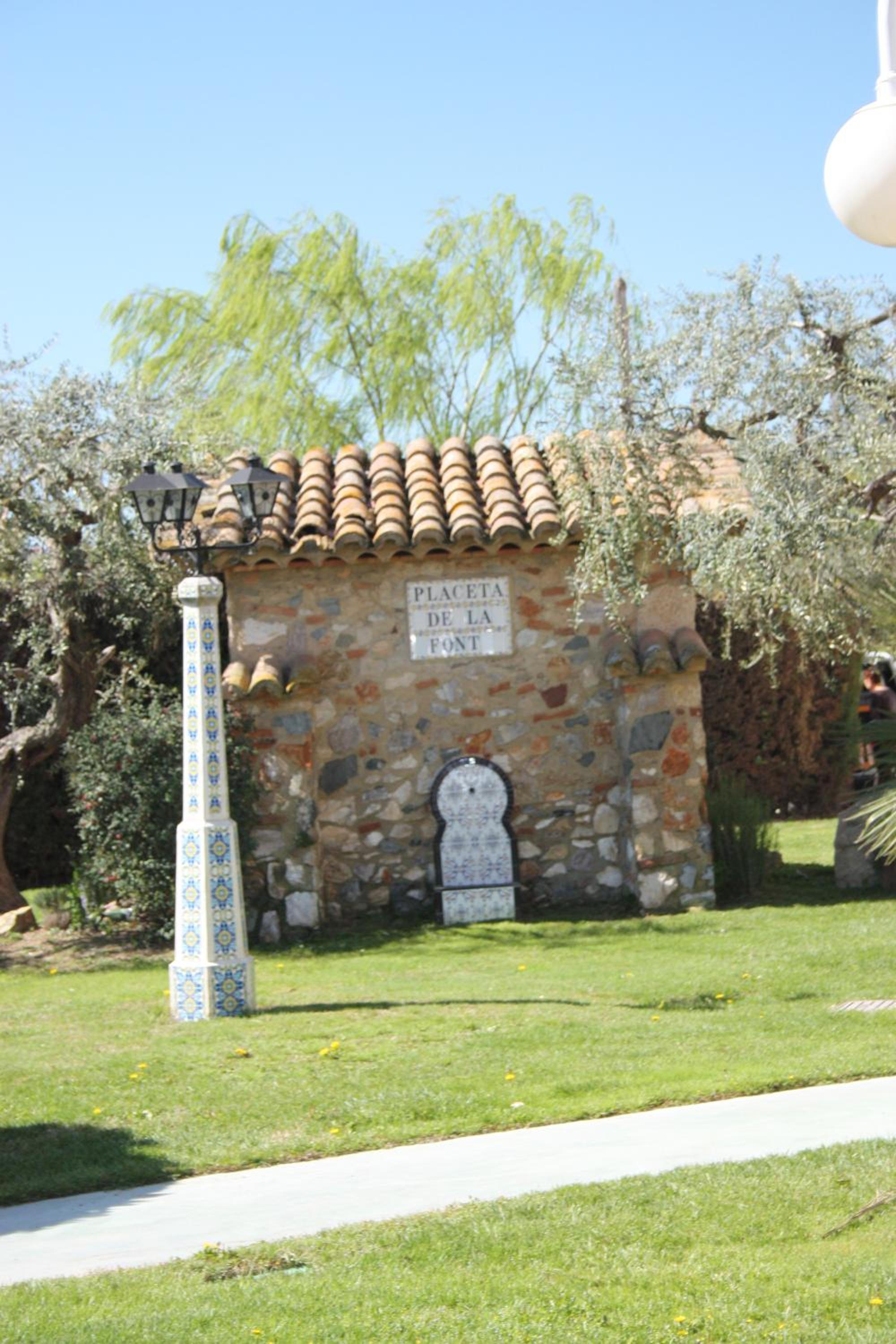 A rustic stone fountain house with clay roof tiles on a grassy lawn beside an ornate tiled lamppost.