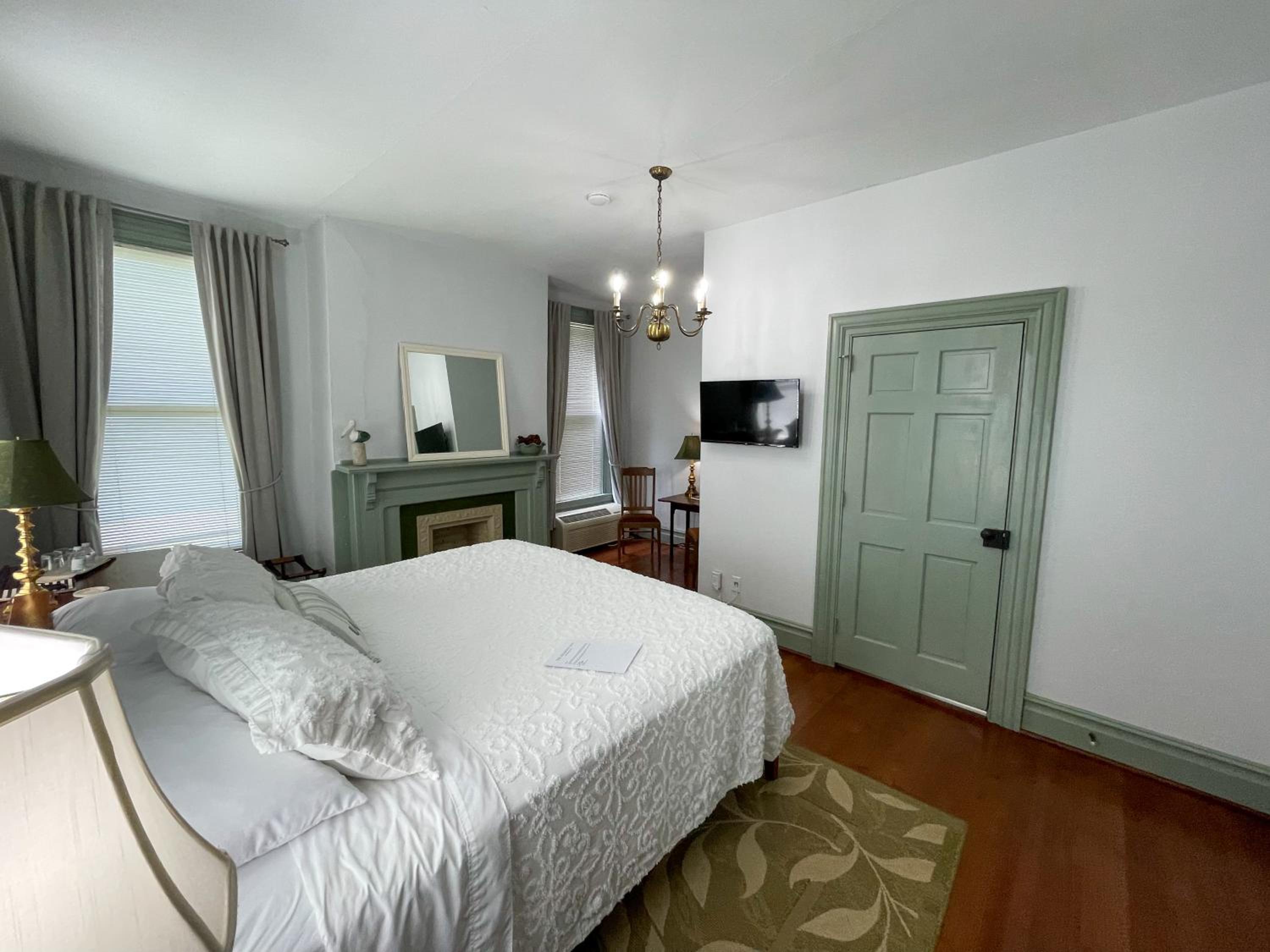 A bedroom with a white-covered bed, sage green trim and fireplace mantel, wood flooring, and a chandelier.