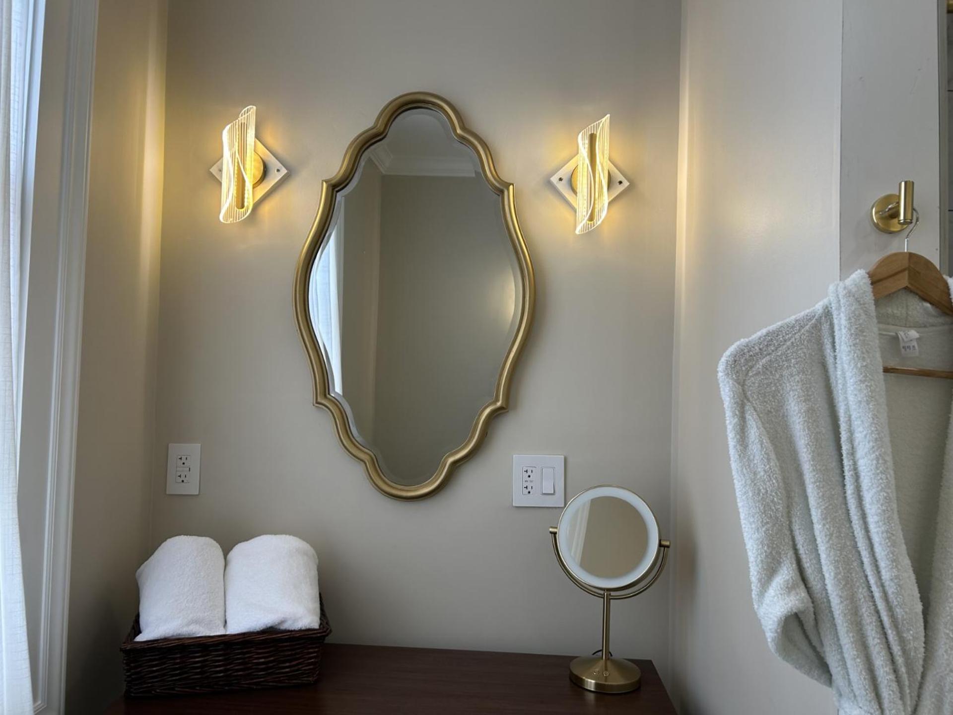 A vanity wall features a gold-framed scalloped mirror flanked by modern sconces, a small mirror, and a white bathrobe.