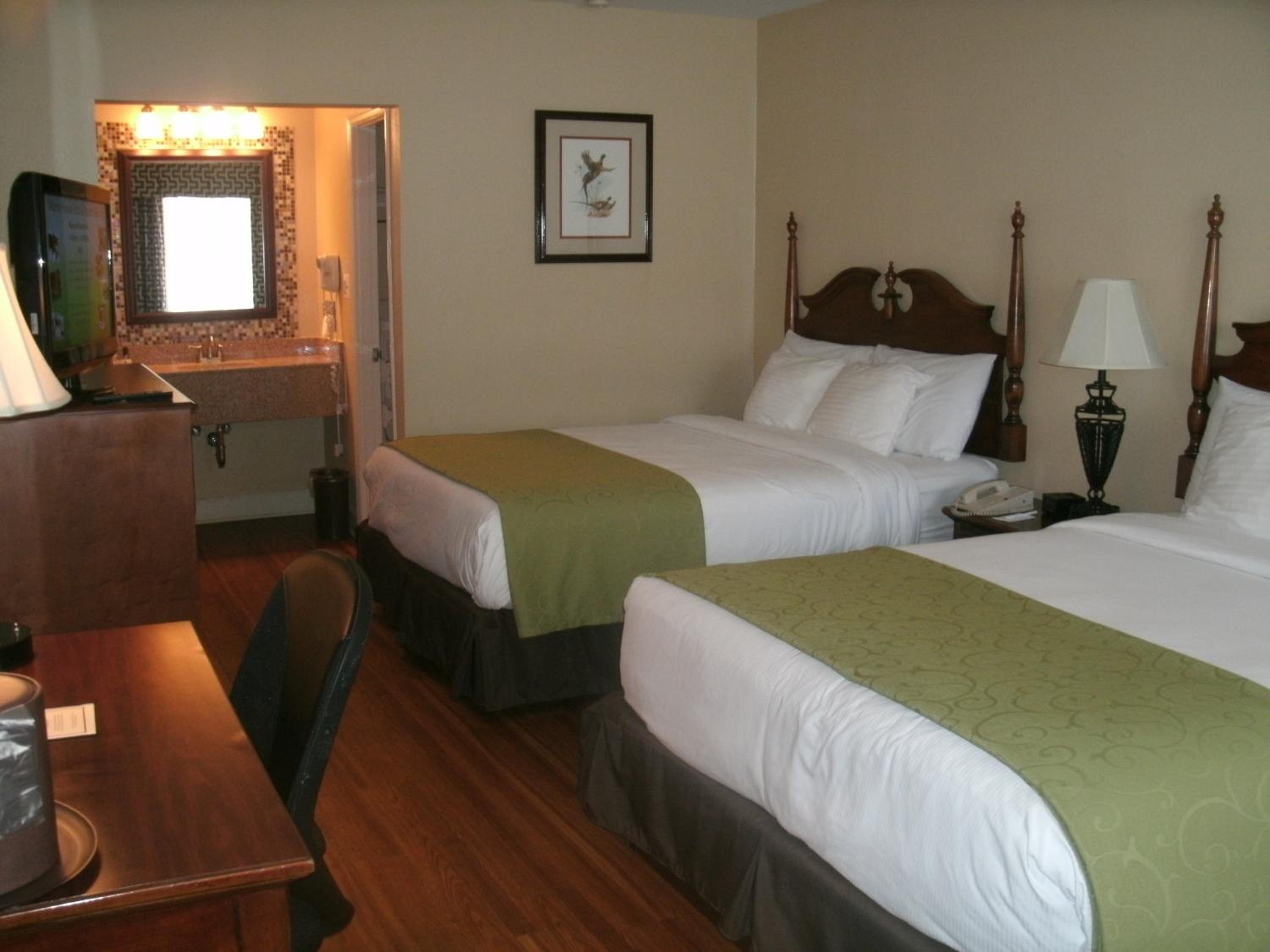 Hotel bedroom with wooden post headboards, green bed runners, a desk, and an adjoining open vanity area.