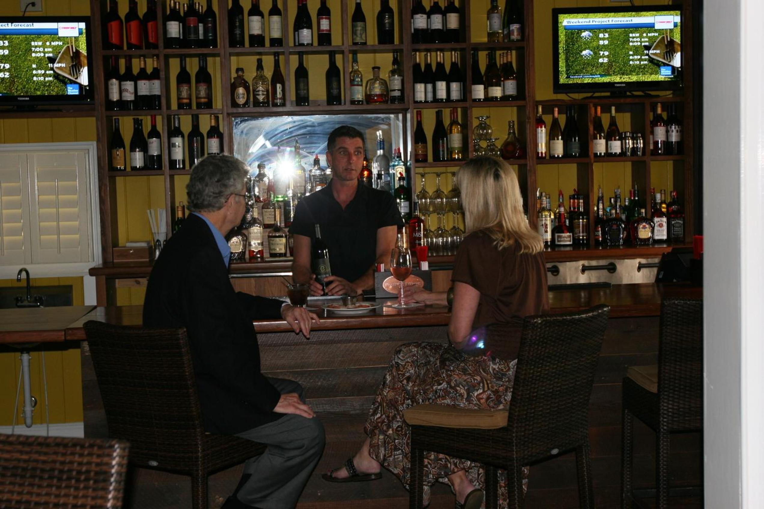 Bartender speaking to patrons seated at a wooden counter backed by shelves of wine and liquor bottles.
