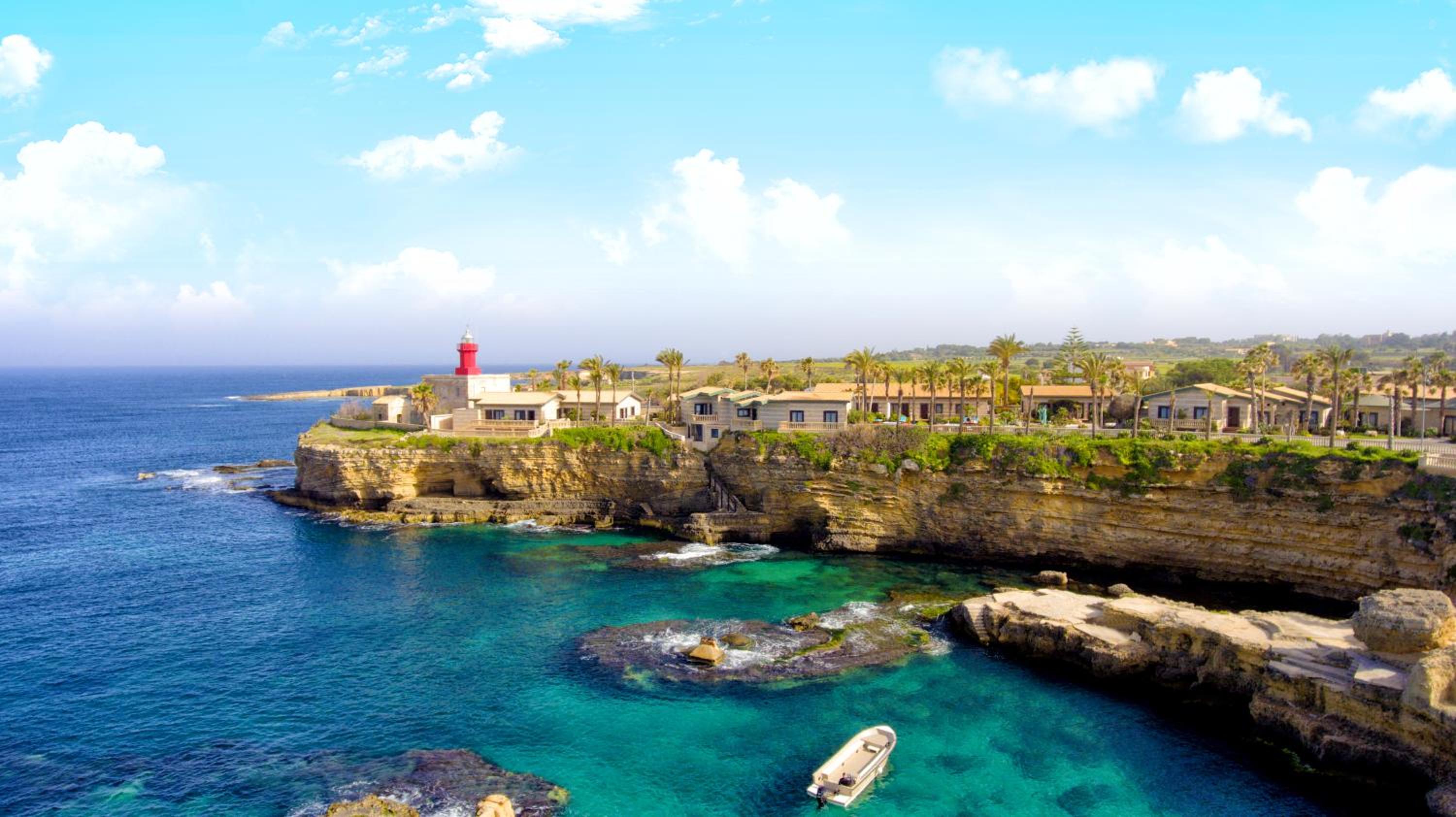 A rocky coastal promontory at Minareto with low stone buildings, a red lighthouse, and clear turquoise sea below.