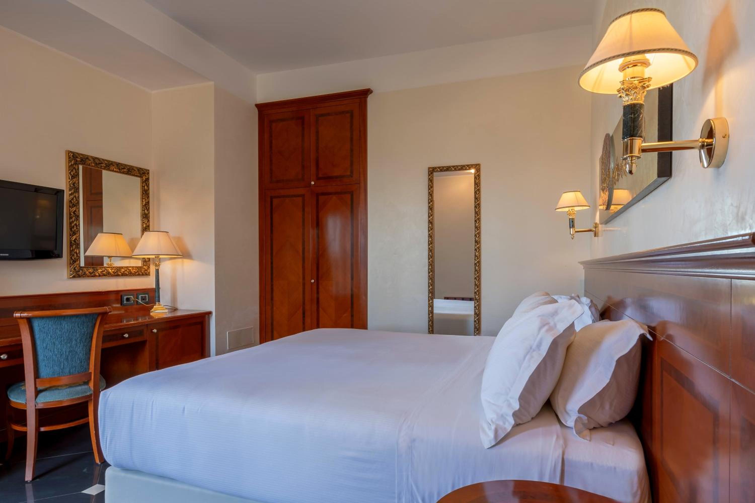 A hotel bedroom with a white-dressed bed, wooden furnishings, warm wall lamps, and a tall mirror.