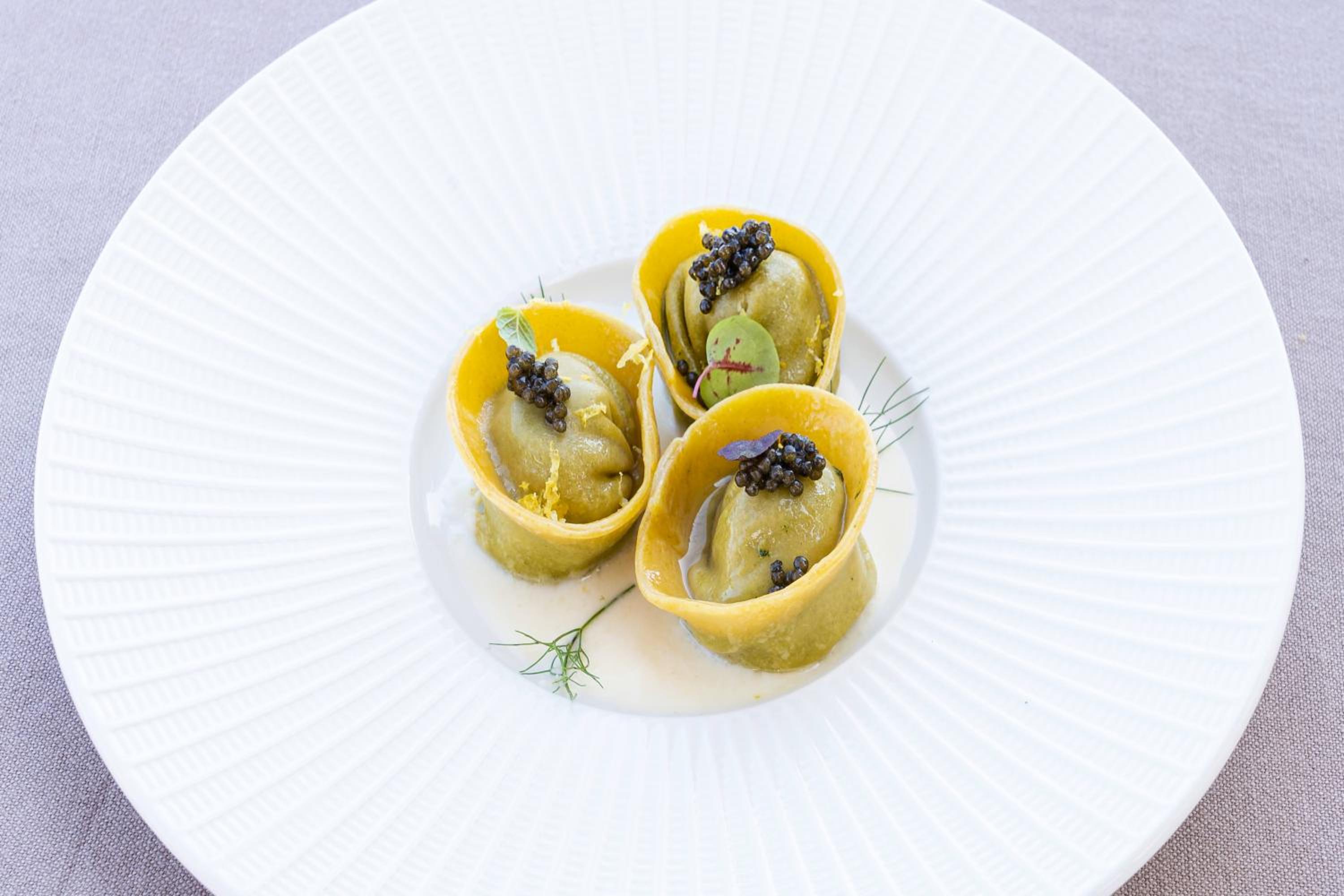 Green filled pasta topped with black caviar and herbs, served in a shallow broth on a white textured plate.