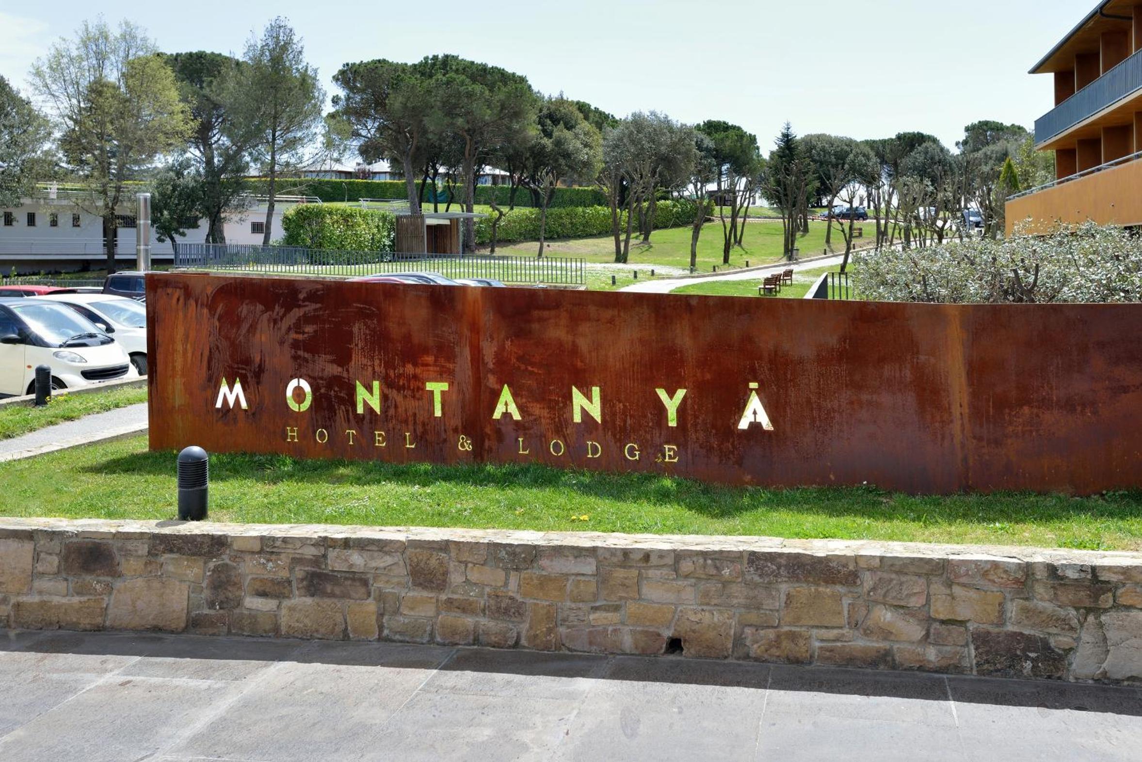 Rusted metal entrance sign reading Montanyà Hotel & Lodge set above a stone wall near a green lawn.