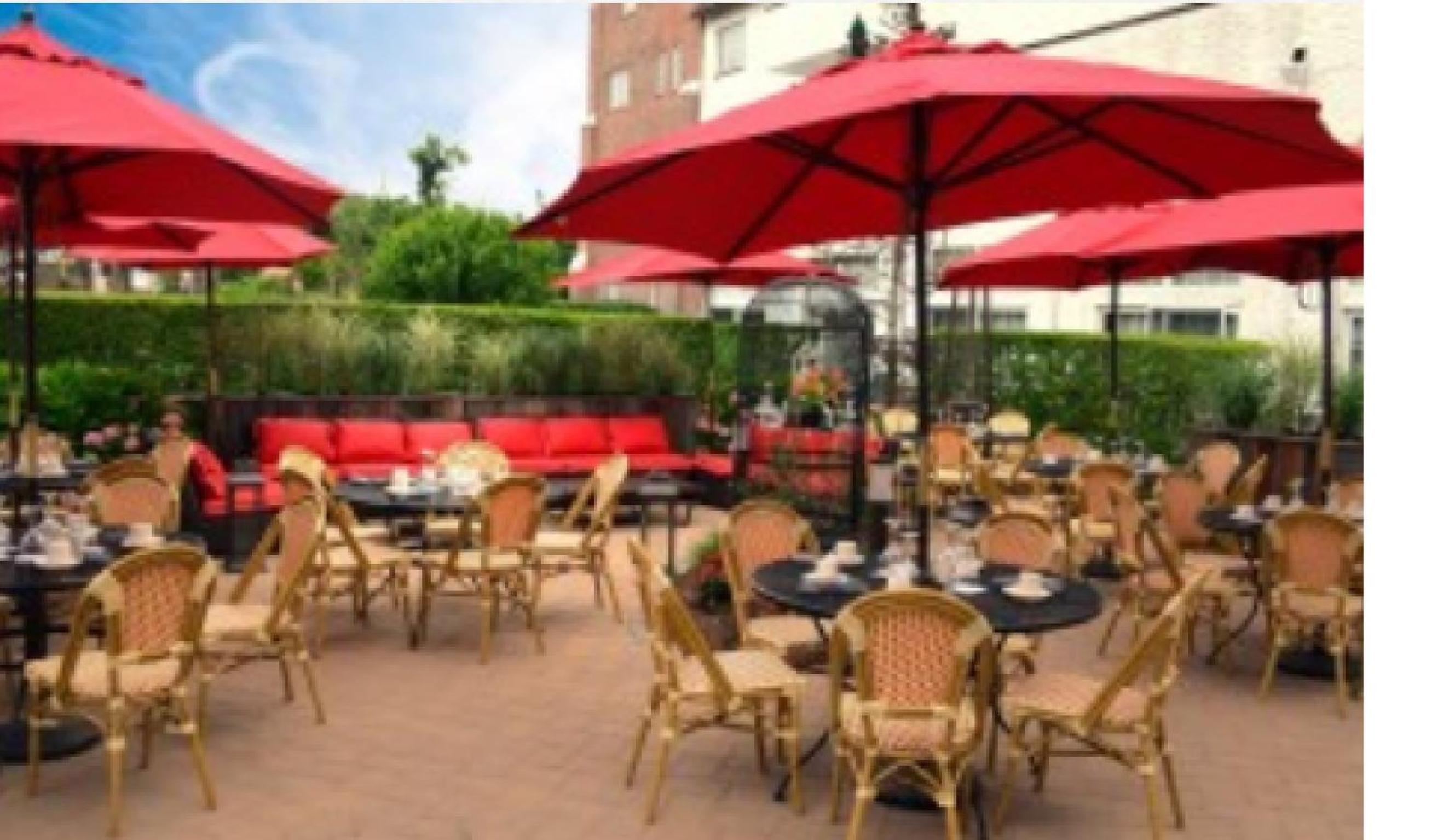 Outdoor brick patio set with dining tables, woven chairs, and red umbrellas beside trimmed hedges.