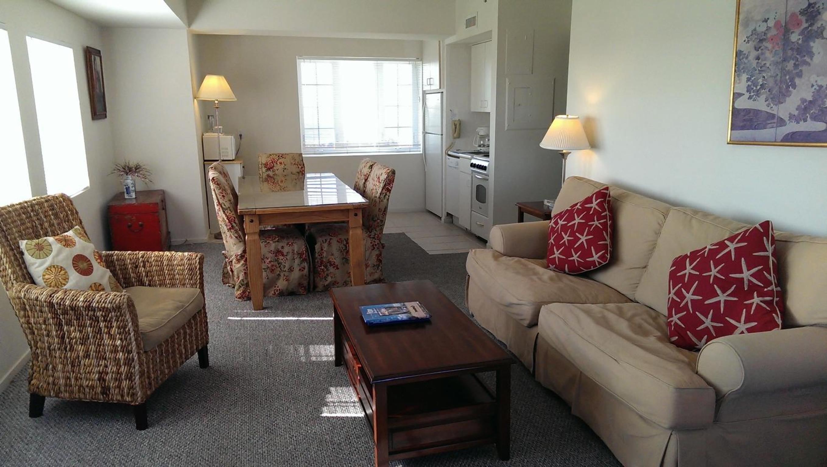 Carpeted living and dining area with neutral sofa, woven armchair, dining set, and an open kitchenette.