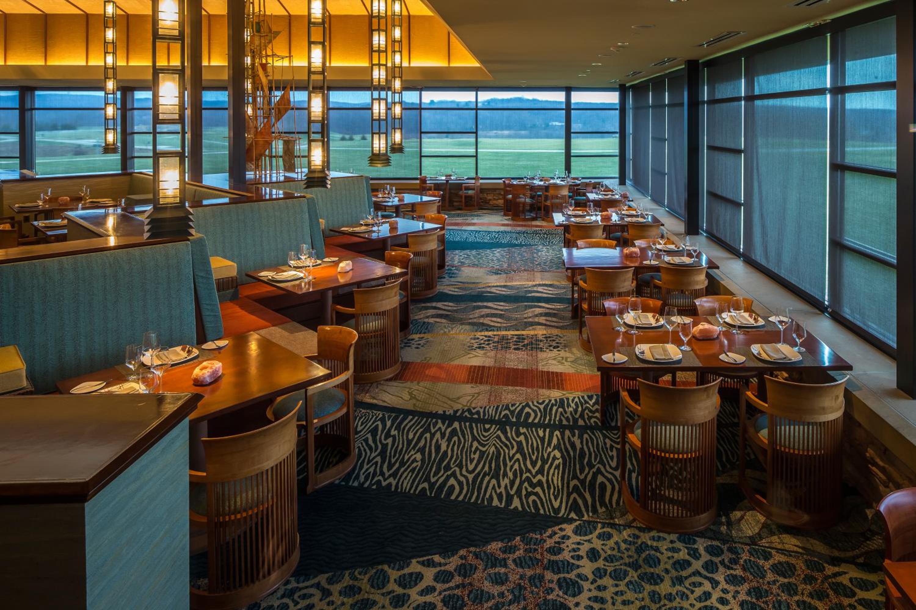 A dining room with wooden tables, patterned carpet, and wide windows looking out onto green fields.