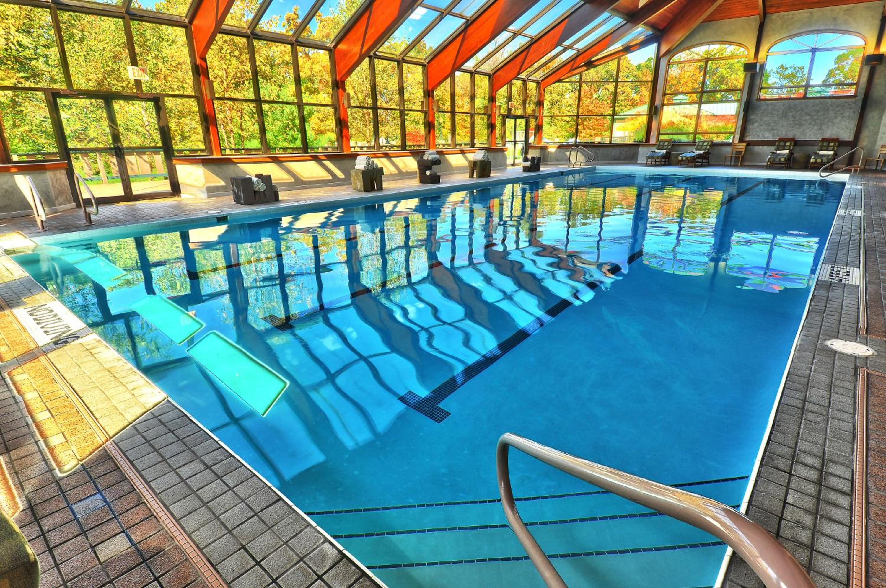 An indoor lap pool surrounded by tile decking under a glass-enclosed timber atrium looking out at autumn trees.
