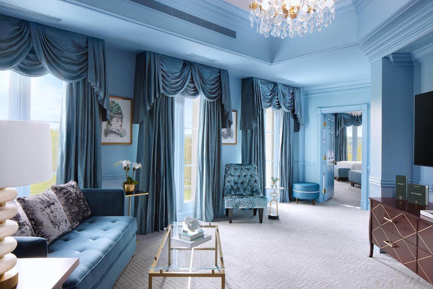 A blue lounge with heavy blue drapes, a velvet sofa, a glass coffee table, and a crystal chandelier.