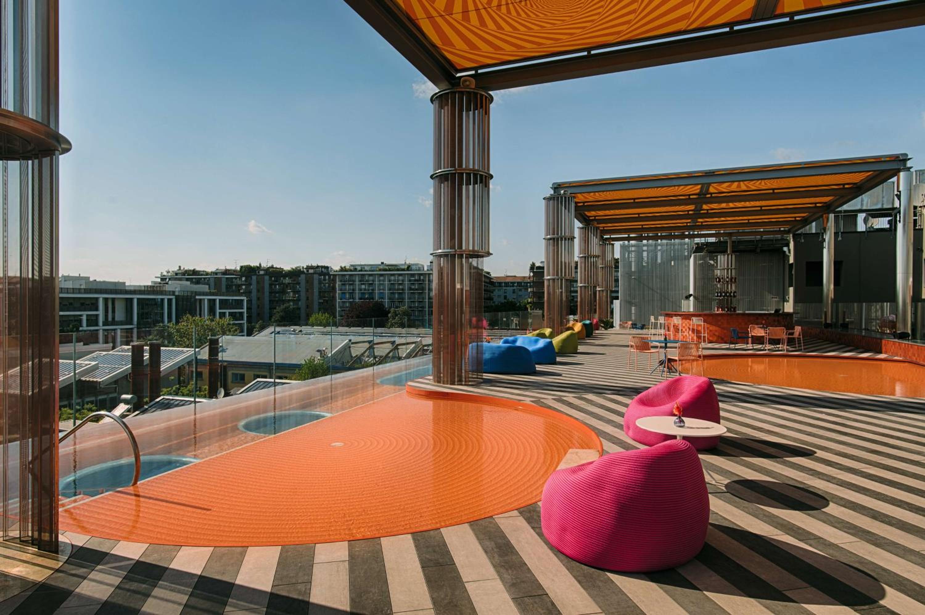 A rooftop terrace with striped decking, vibrant beanbag seats, a shallow orange pool, and city views.