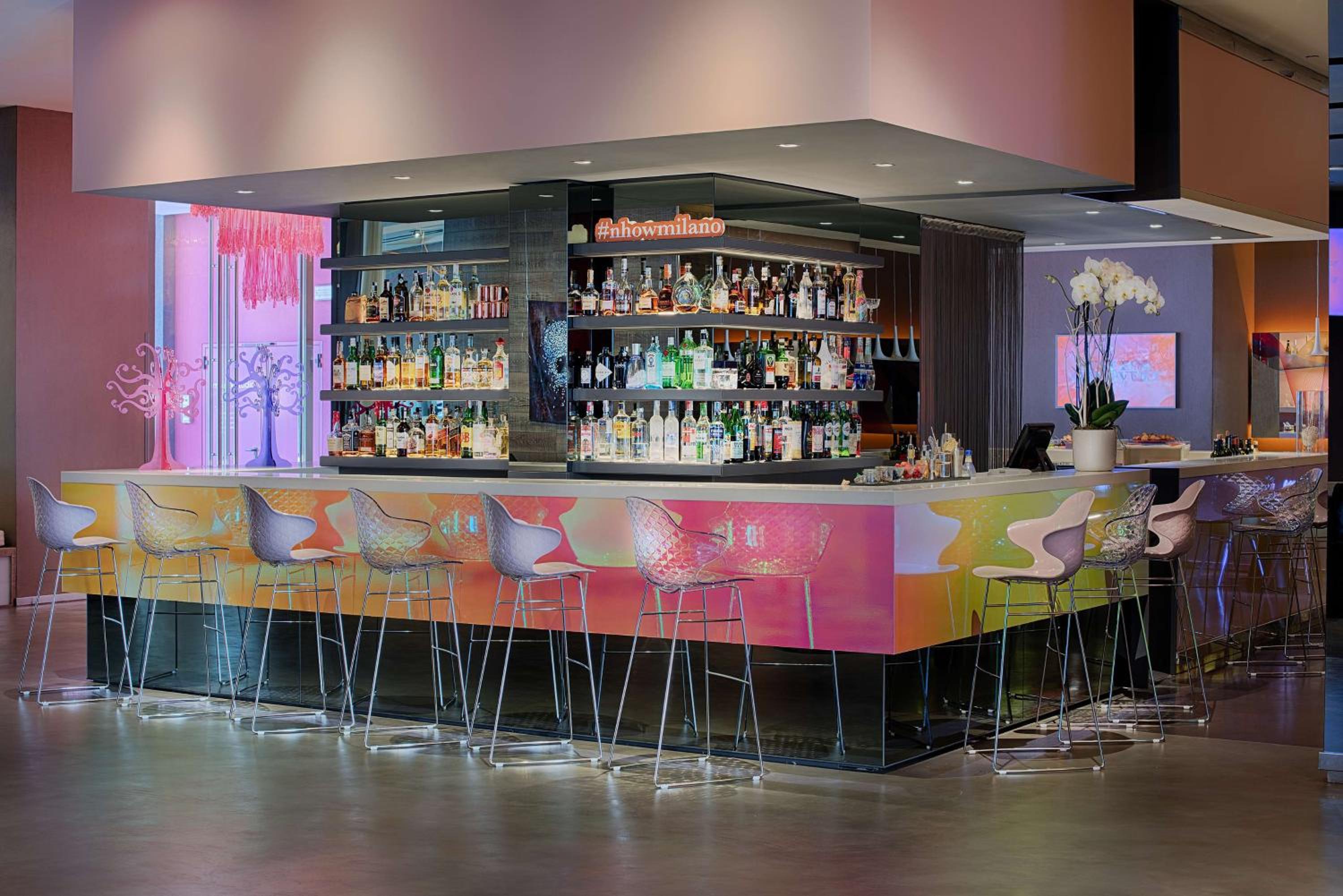 A contemporary bar with an iridescent front panel, high barstools, and illuminated shelves of bottles.