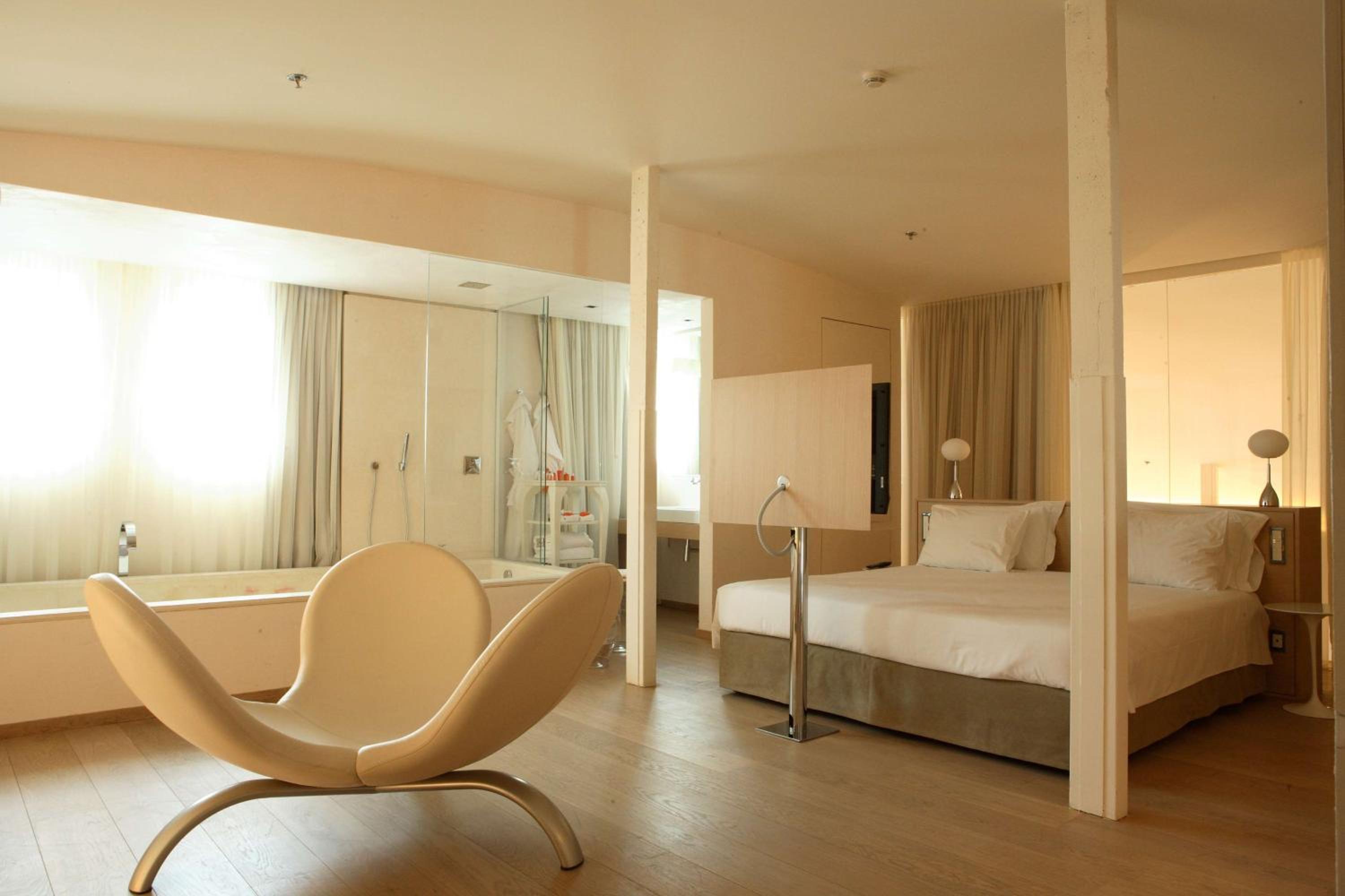 An open-concept suite with a bed, light wood flooring, an accent chair, and a glass-walled bathroom.
