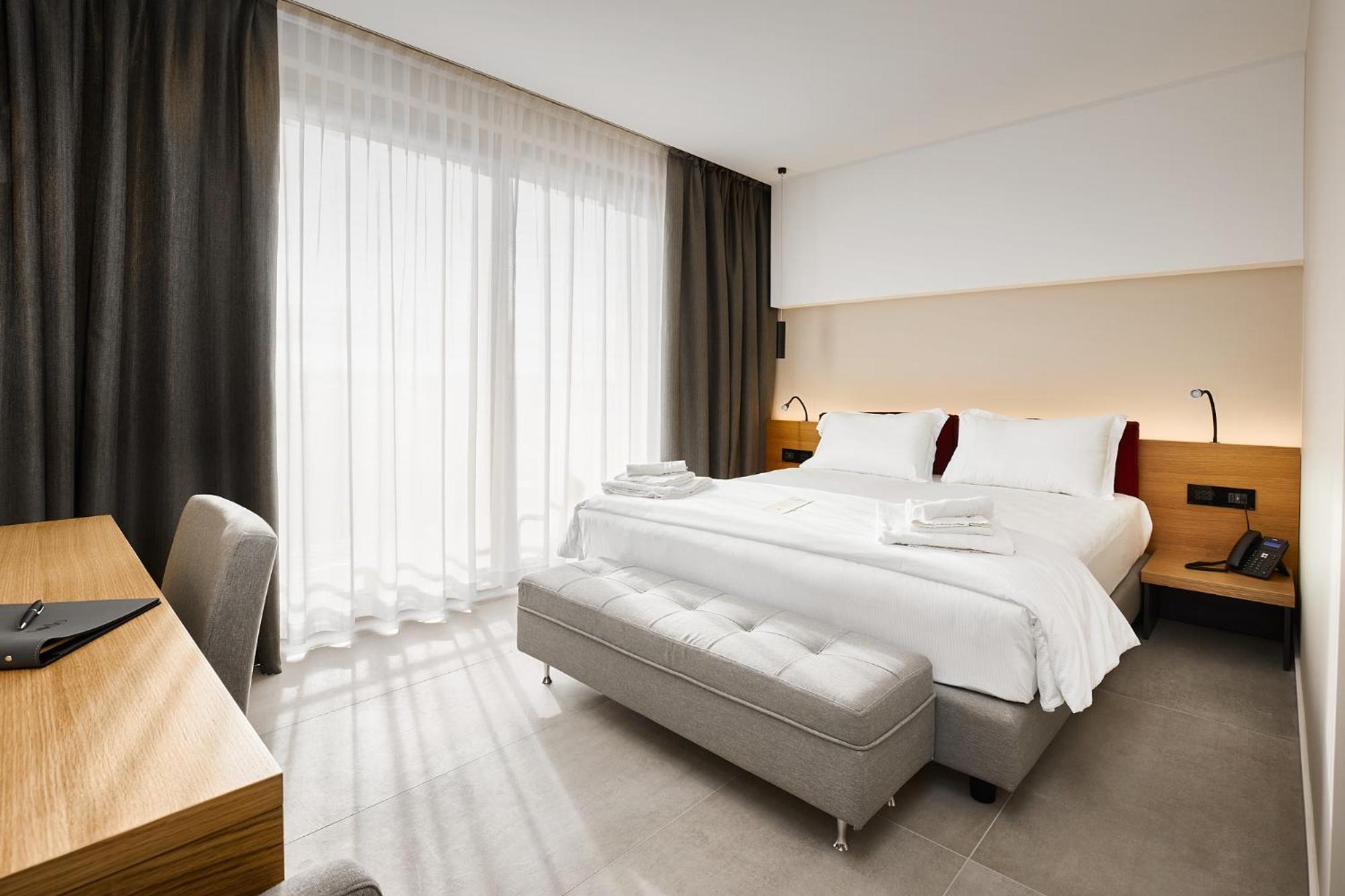 A bright hotel bedroom with a bed, wooden headboard, and sheer curtains filtering sunlight.