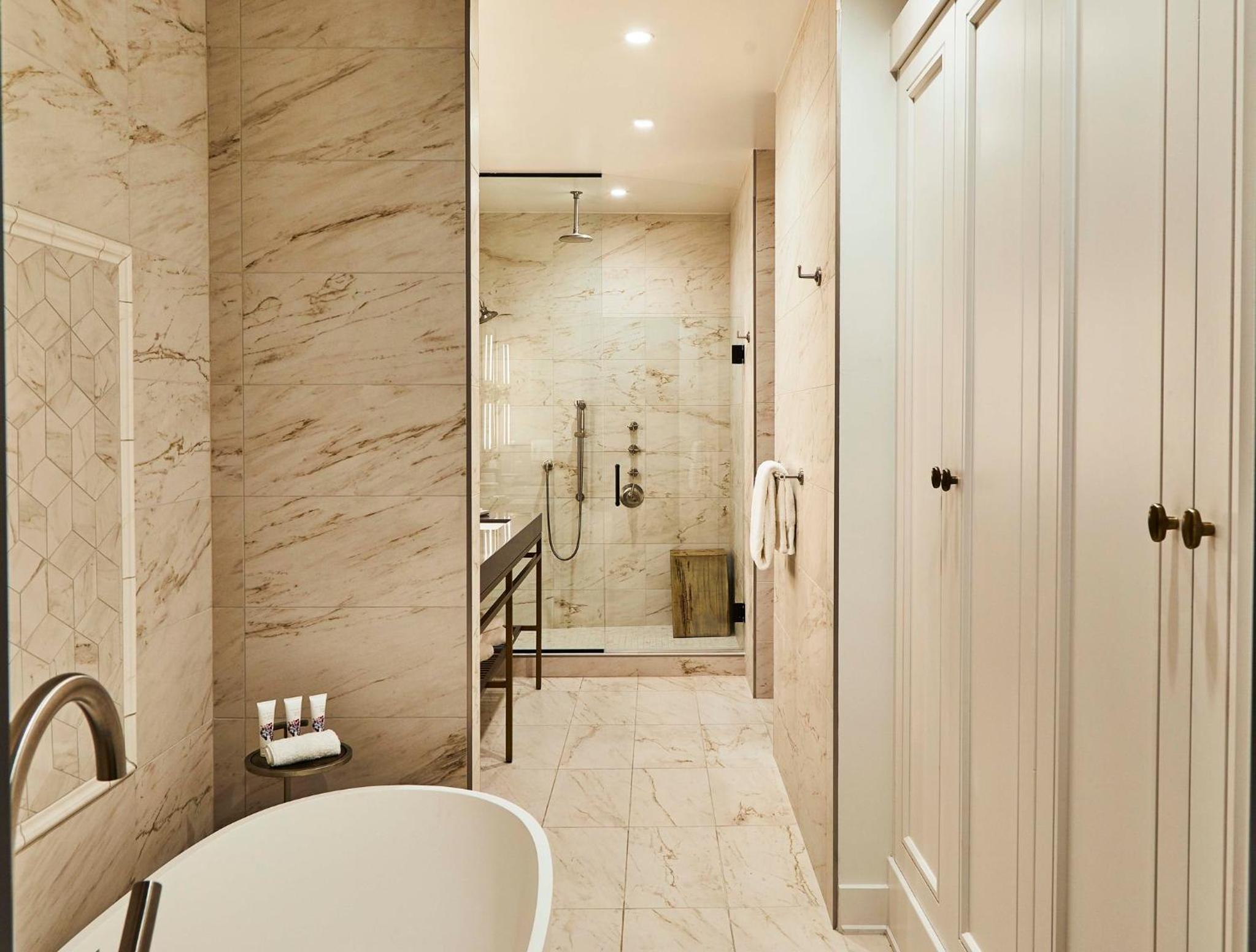 A marble-tiled bathroom with a freestanding soaking tub, glass walk-in shower, and tall white paneled cabinets.