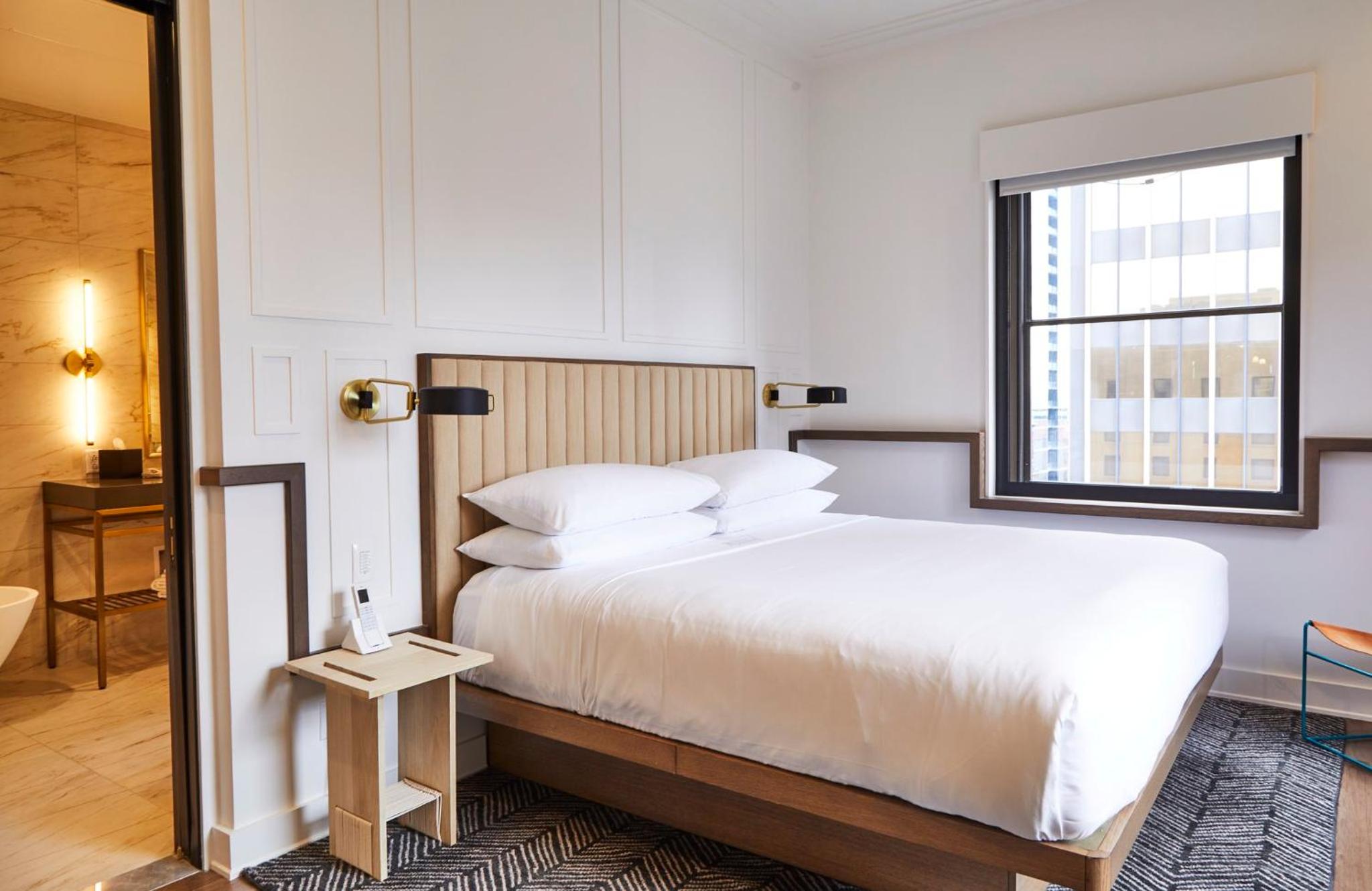 A hotel bedroom with a wood-framed bed, fluted headboard, geometric rug, and a window framing city buildings.