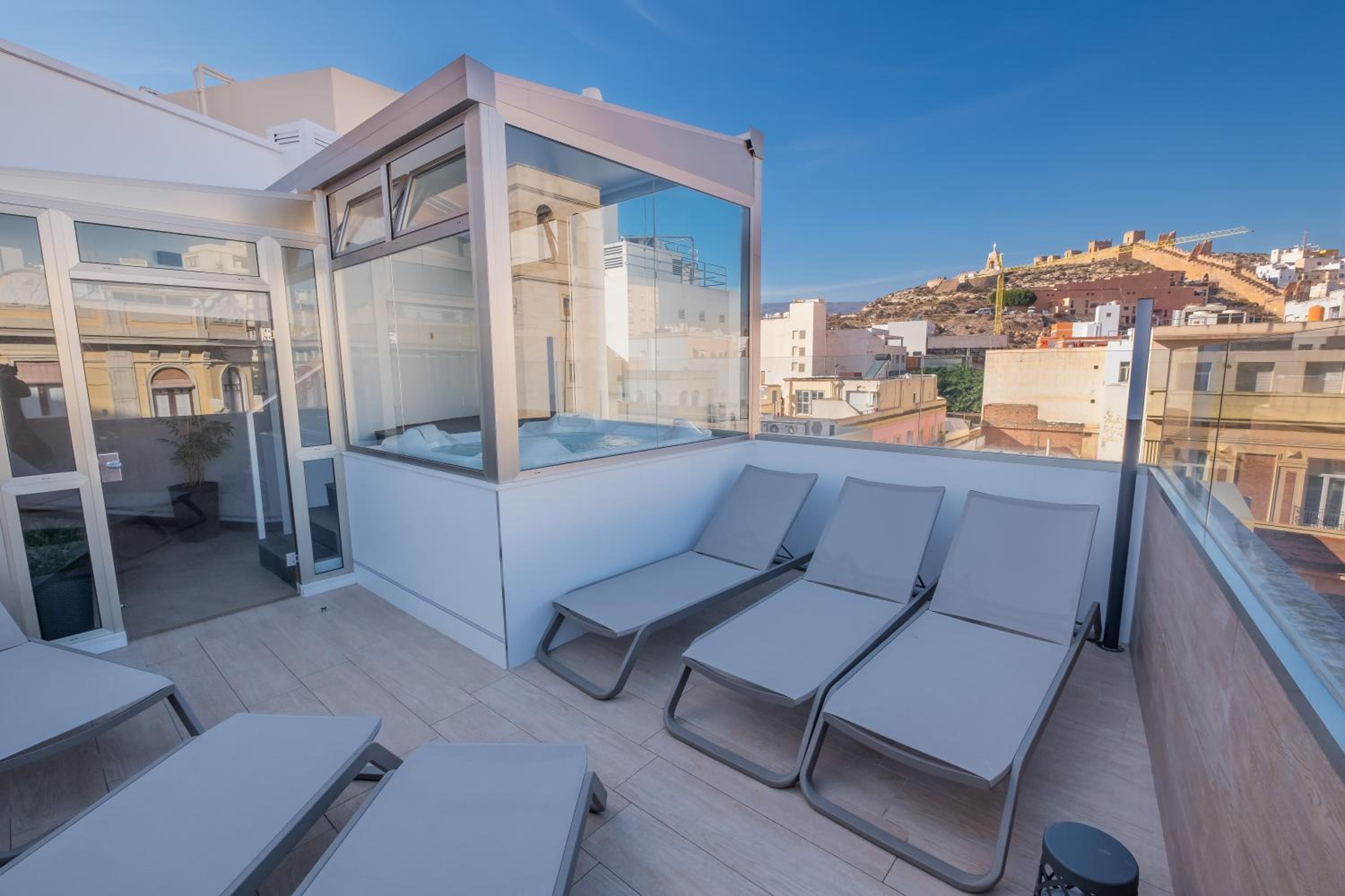 Rooftop terrace with grey sun loungers, an enclosed glass hot tub, and a view of an ancient fortress on a hill.