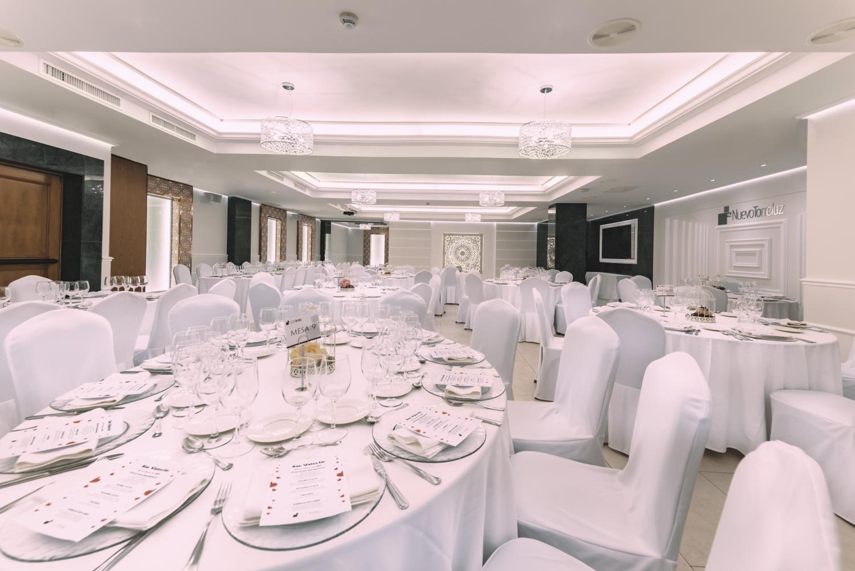 Banquet hall at Nuevo Torreluz set with round tables, white tablecloths, crystal chandeliers, and white chair covers.