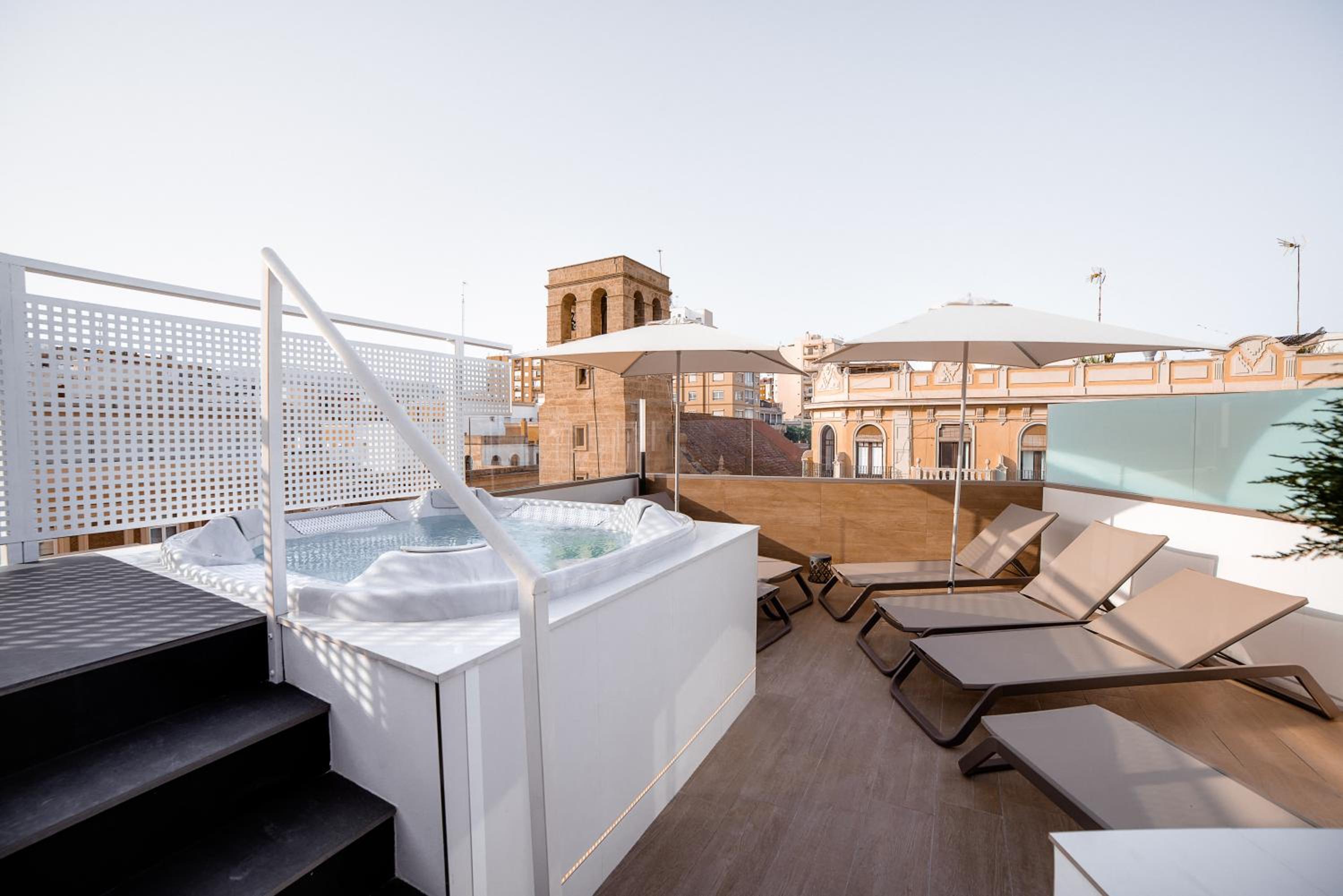 Rooftop terrace featuring an outdoor hot tub, sun loungers under white parasols, and surrounding city buildings.
