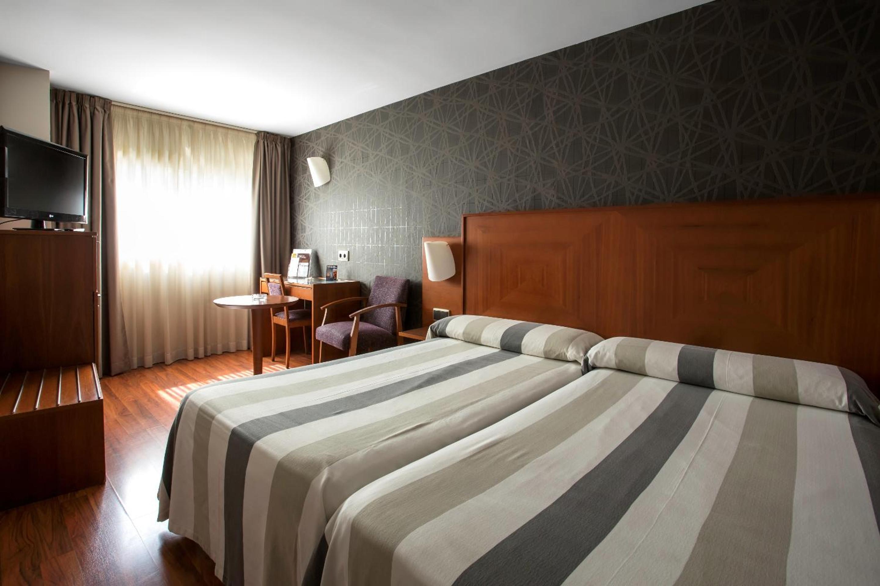 Hotel bedroom with wooden headboard, striped bedspreads, patterned wallpaper, a desk, and a television.