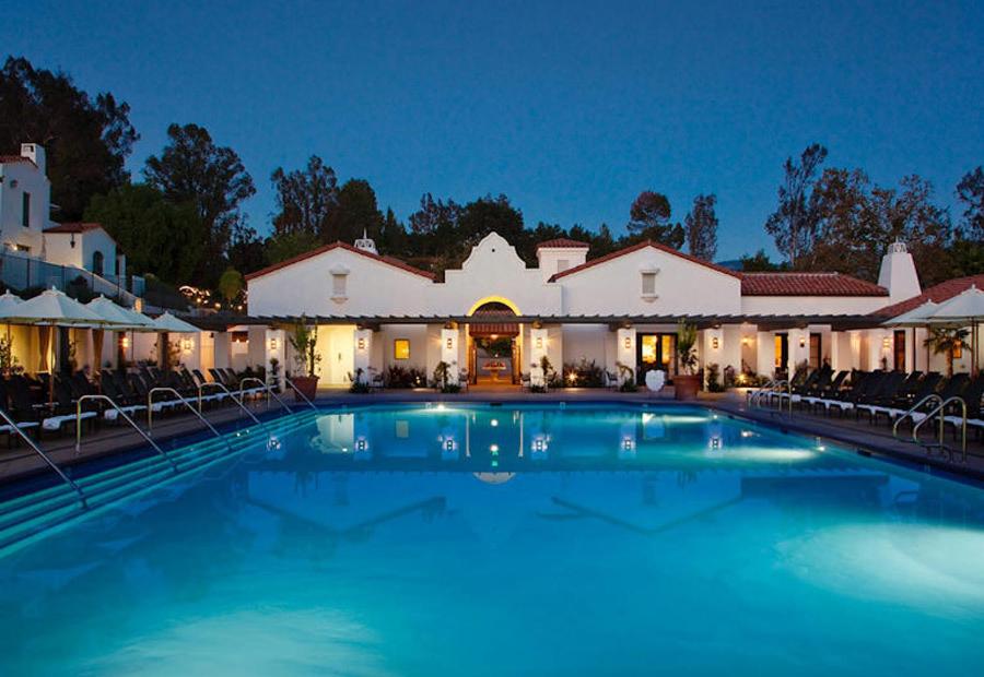 A swimming pool illuminated in blue at dusk, bordered by lounge chairs and white tile-roofed villas.