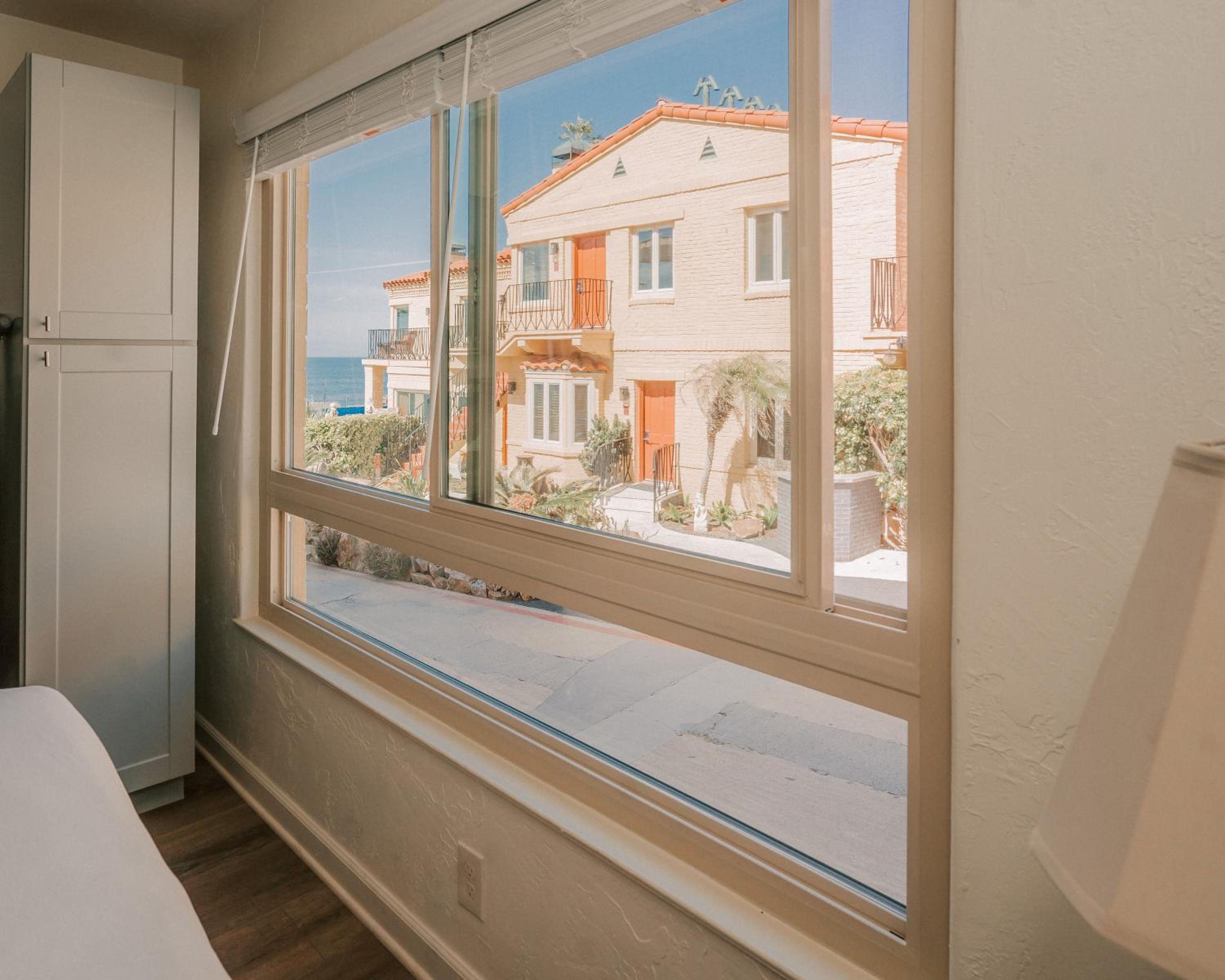A view through a window looks out at tan stucco buildings and the blue ocean beyond.