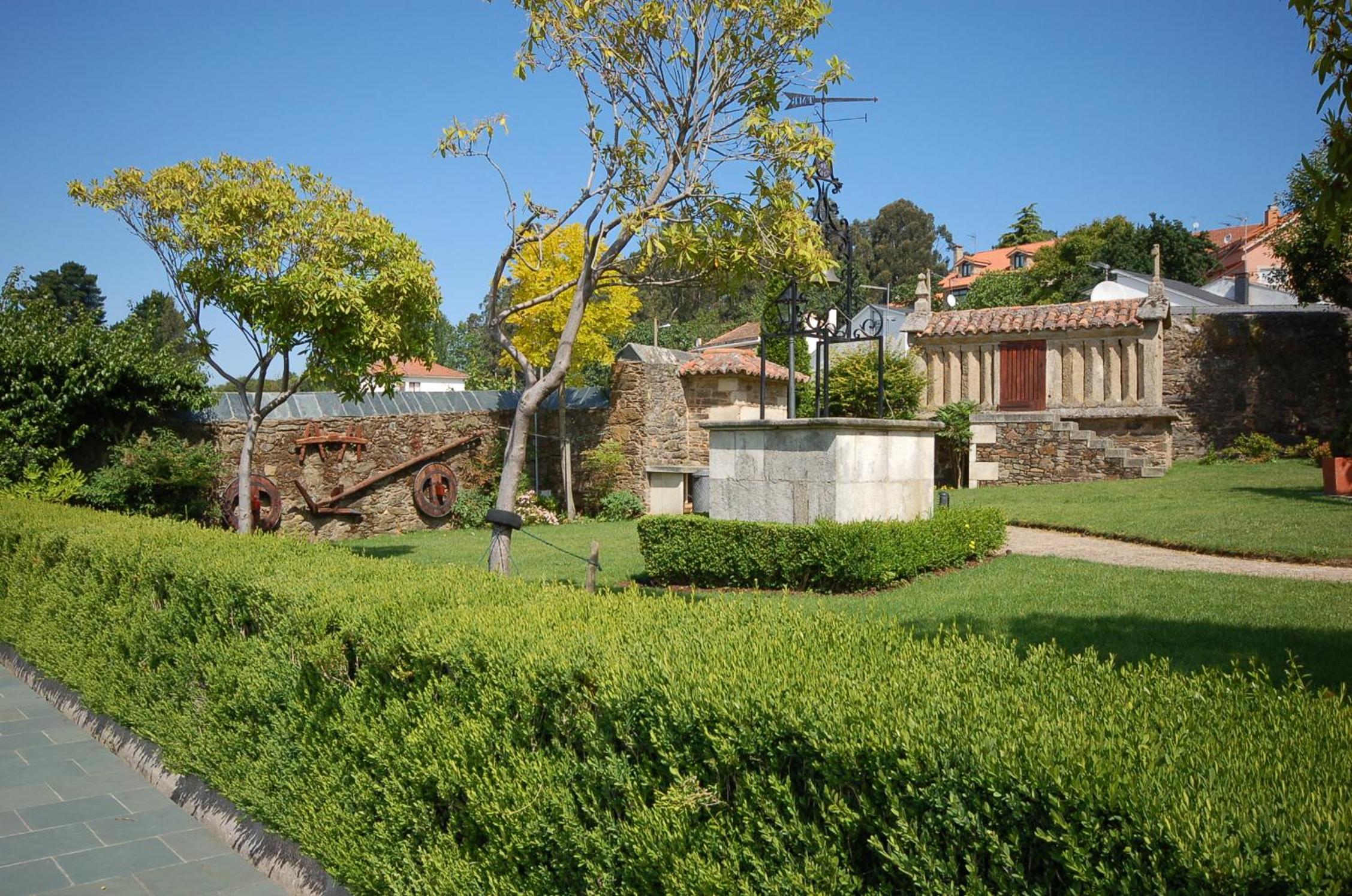 Stone well and traditional Galician granary in a grassy garden bordered by a stone wall and hedges.