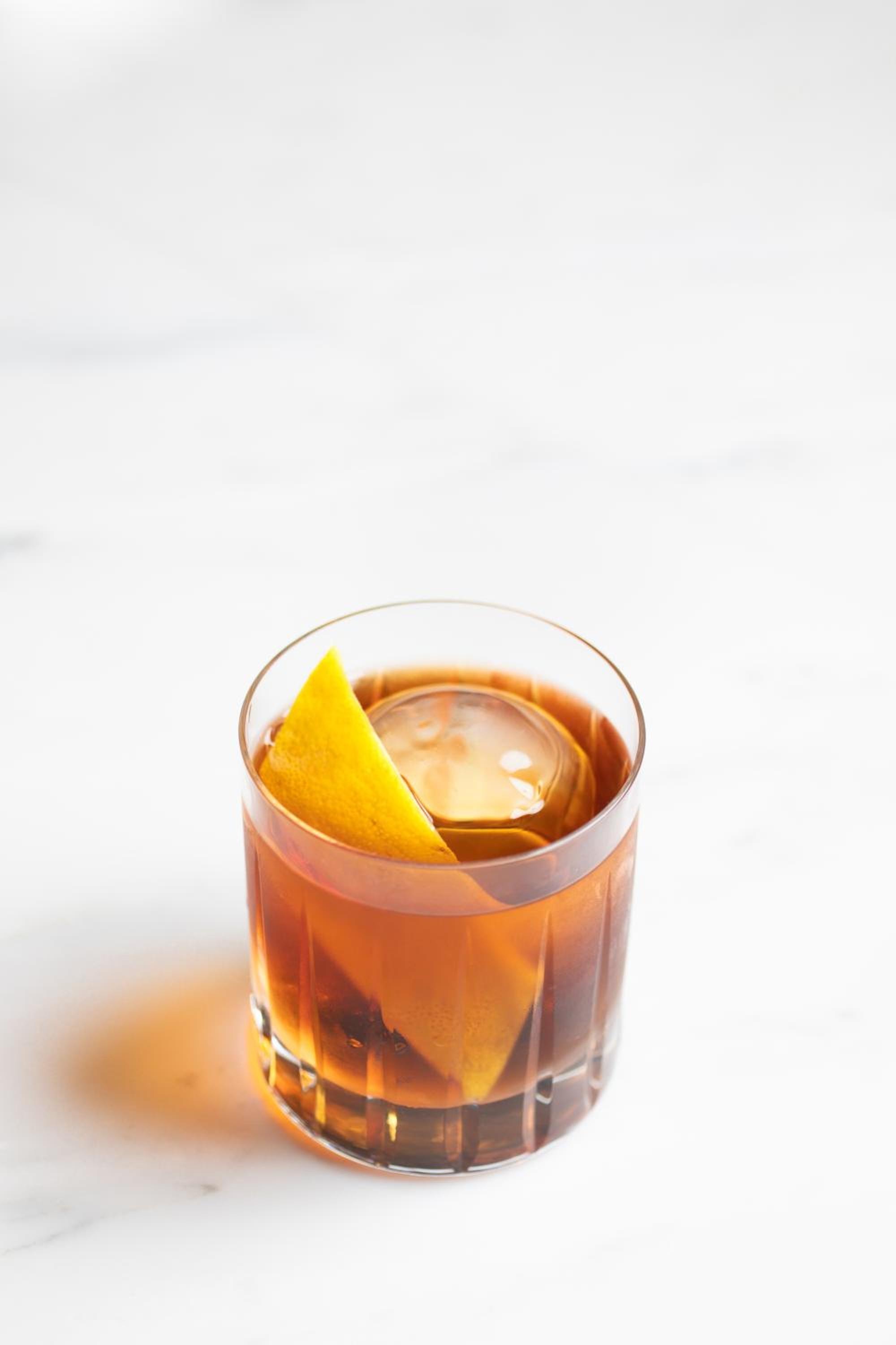 An amber cocktail served over a round ice sphere with citrus peel on white marble.
