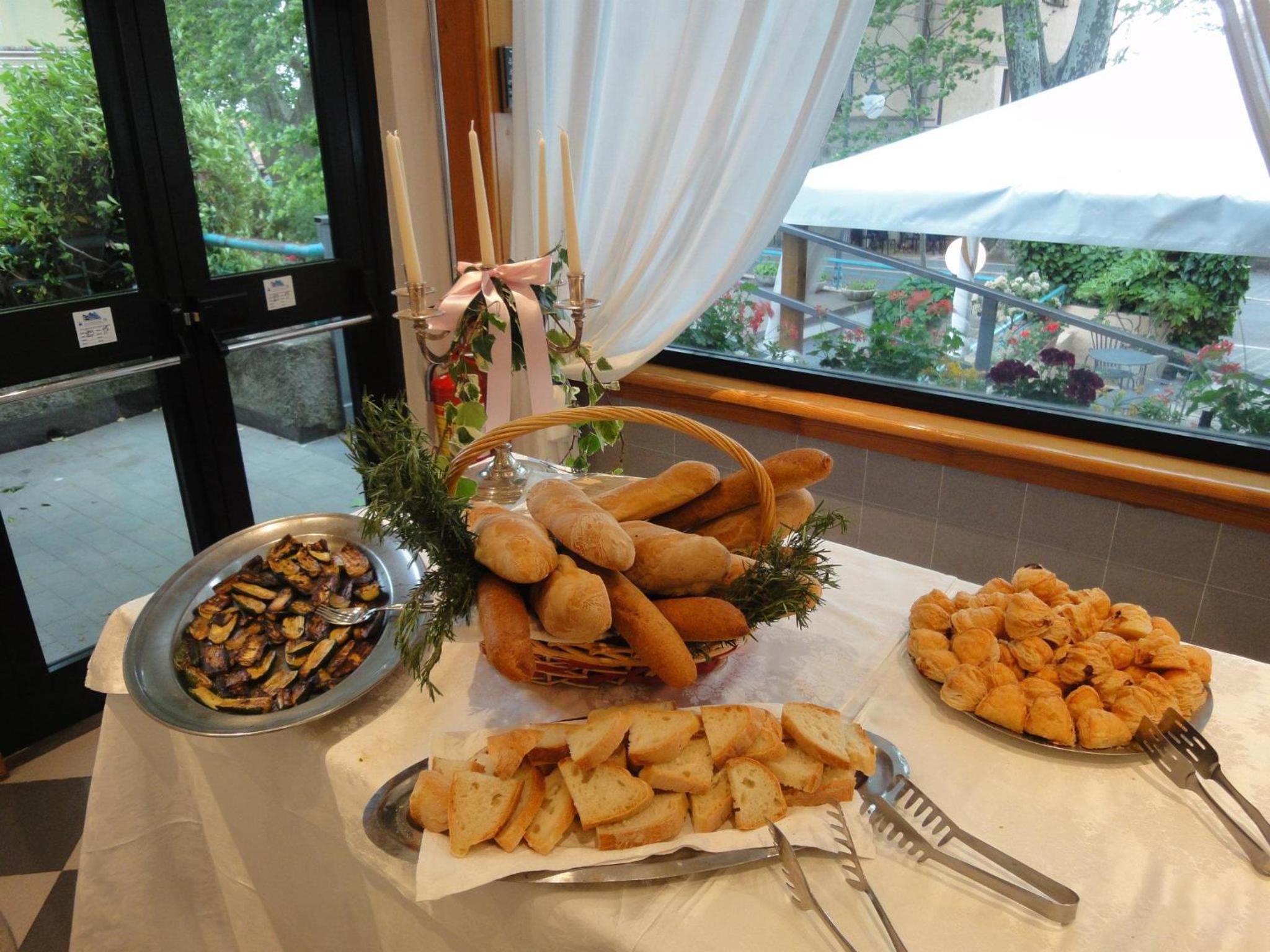 Buffet table set with baskets of sliced bread, grilled vegetables, and pastries near a large window with candelabra.