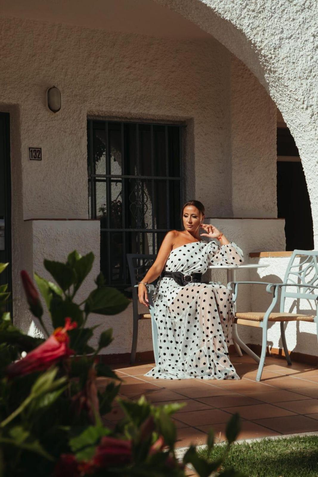 A woman in a polka-dot dress rests on an outdoor patio chair beside a white textured wall and potted plants.
