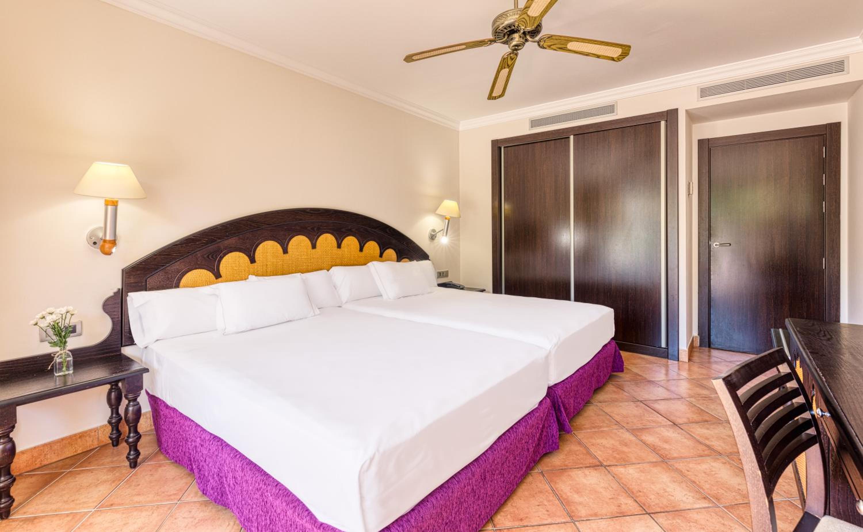 A hotel bedroom with tiled floors, purple-skirted beds, an arched wooden headboard, and dark wood wardrobes.