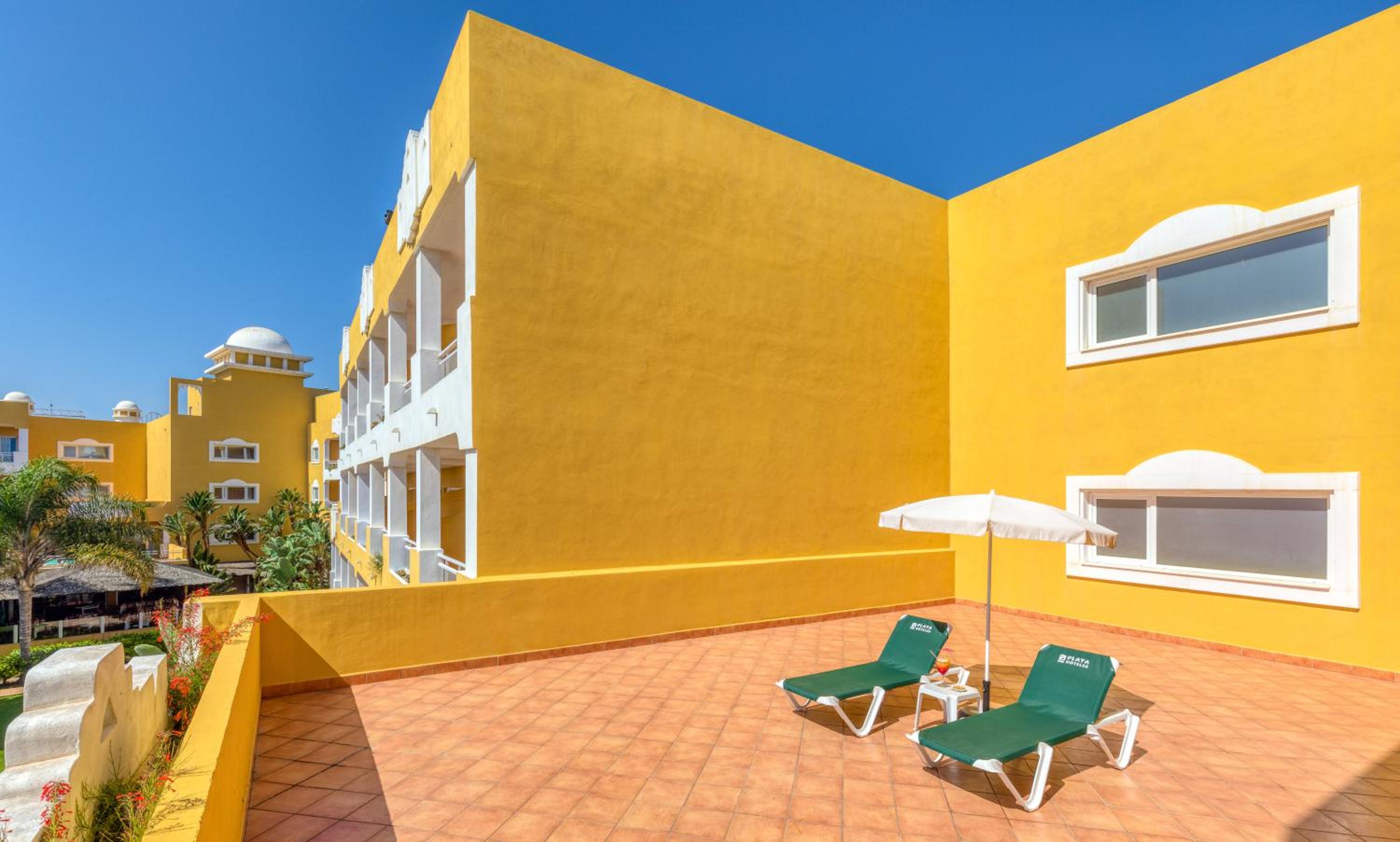 A tiled rooftop terrace with green loungers and a white parasol against bright yellow resort walls.