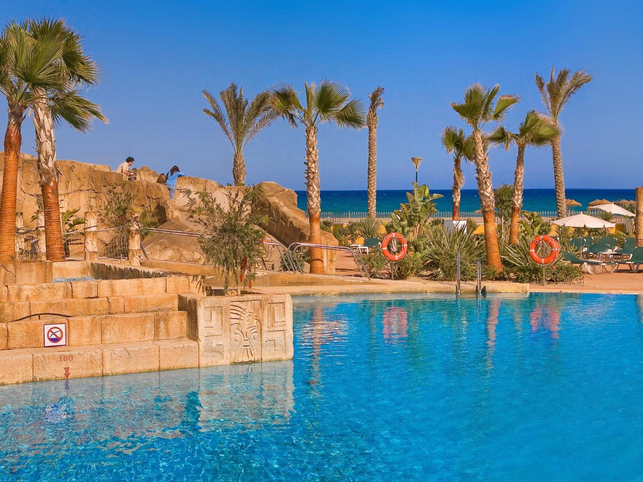 A resort swimming pool with carved stone blocks, palm trees, and the blue sea visible in the background.