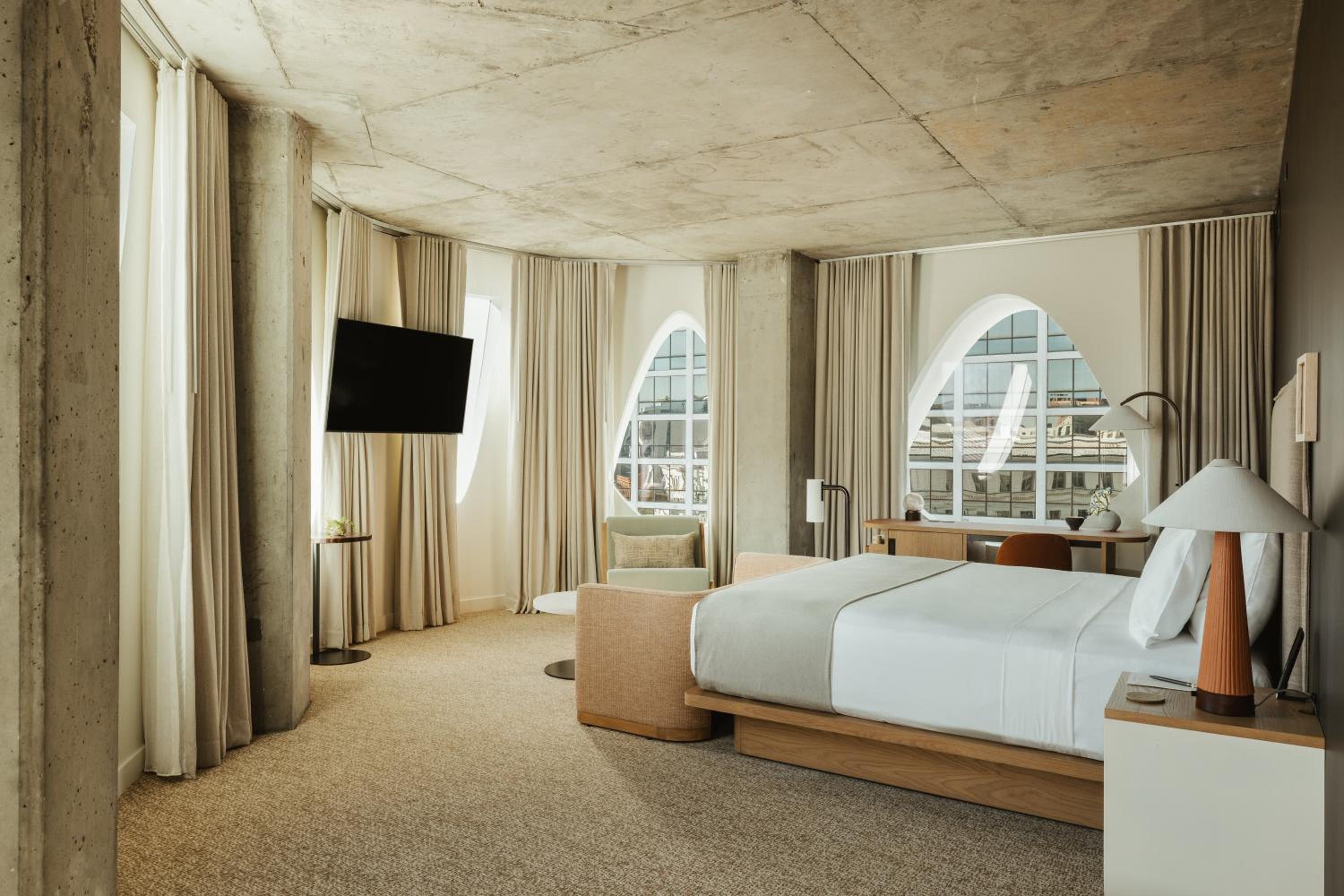 A hotel bedroom with raw concrete ceilings, arched windows, light drapery, and a low platform bed.